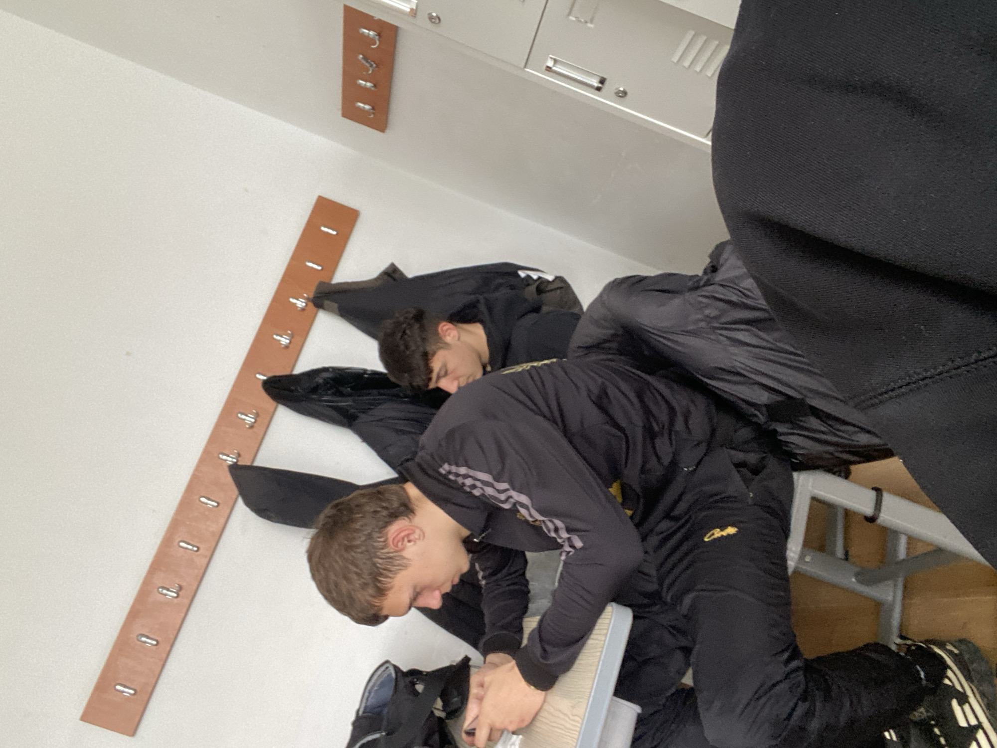 Two young men are in what appears to be a locker room with metal lockers and a wooden coat rack attached to the wall. One man is sitting on a stool wearing a black tracksuit, while the other stands behind him. The location is presumably in Varna, Bulgaria.