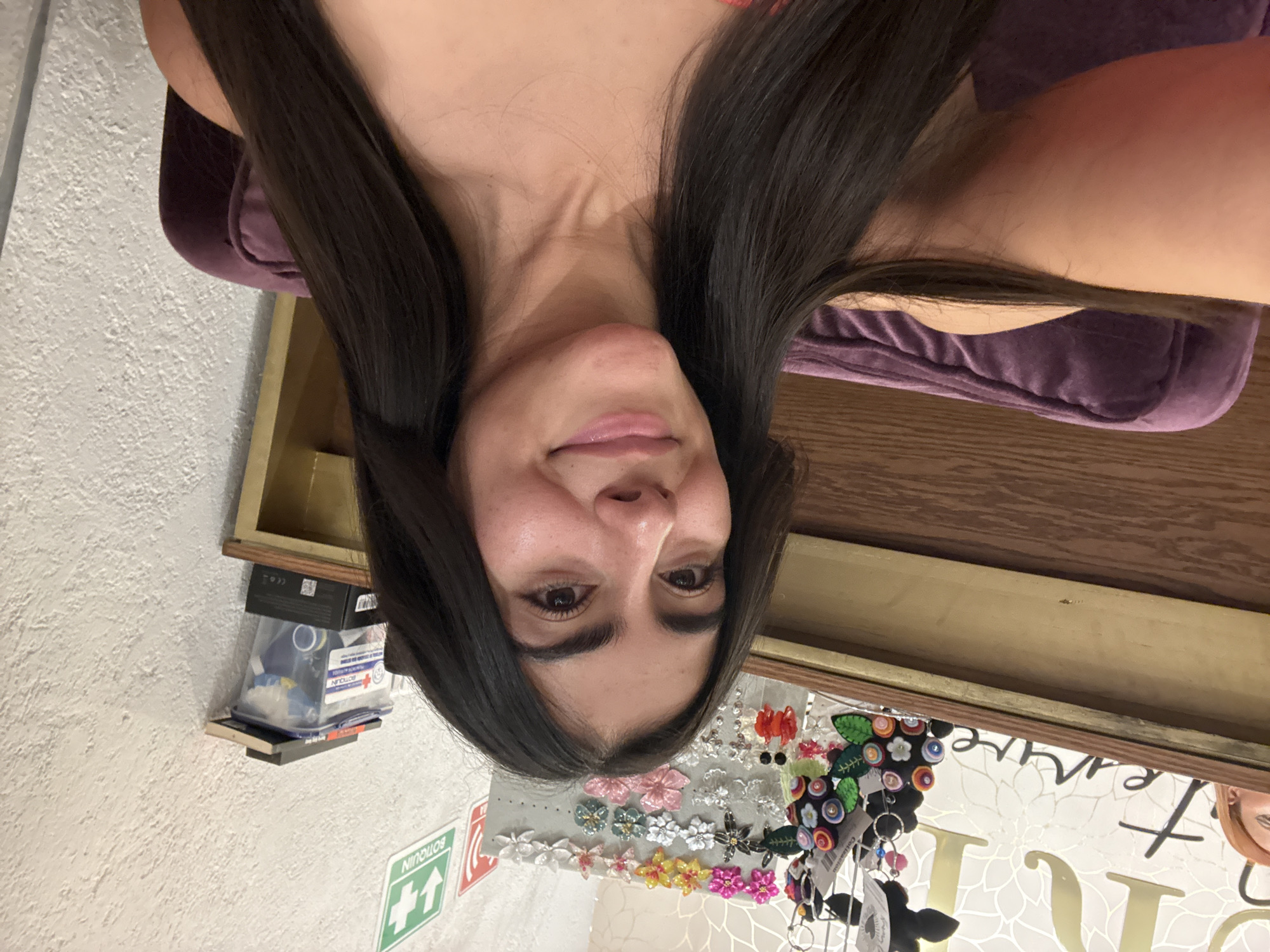 The image shows a woman with long, dark hair, fair skin, and dark eyes. She is facing the camera with a slight smile. The background suggests she's inside a store or small boutique. On the wall behind her, there are displays of hair accessories, including flower pins and keychains. A first aid kit is visible mounted on the wall. Text is reflected in the mirror and a partial reflection of another person is visible in the mirror. The location is likely in the city of Domingo Arenas, Mexico.