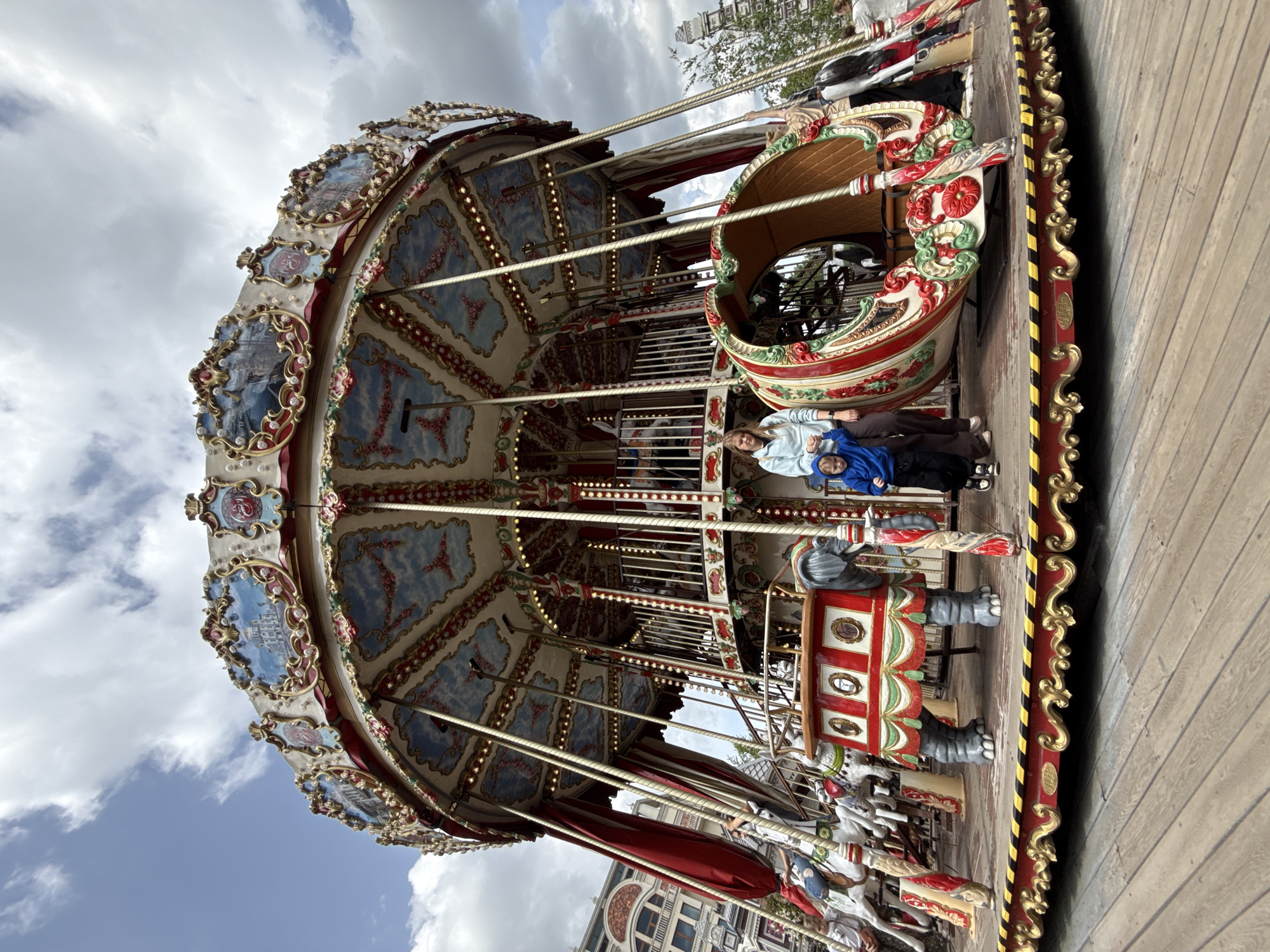 The image depicts a carousel in an outdoor setting. The carousel is elaborately decorated with intricate carvings, gold accents, and colorful paintings of scenic landscapes on its upper section. Red curtains hang from the sides, framing the carousel. In the foreground, there are several carousel figures, including a grey elephant with a decorative red saddle and what appear to be horses.
Two people are visible on the carousel platform. A woman in a light blue hooded jacket and dark pants stands next to a young boy wearing a blue hooded sweatshirt and dark pants. They are standing near the elephant figure. Other figures and parts of the carousel are visible in the background. The carousel is situated on a wooden platform, which appears to be part of a larger public space or amusement area. Buildings with architectural details suggesting a European city can be seen in the background, as well as trees and a cloudy sky. The overall impression is of a vibrant and festive amusement park setting.
The carousel is located at the Moscow Carousel at VDNKh (Exhibition of Achievements of National Economy) in Moscow, Russia. This carousel is known for its historical significance and ornate design, making it a popular attraction.