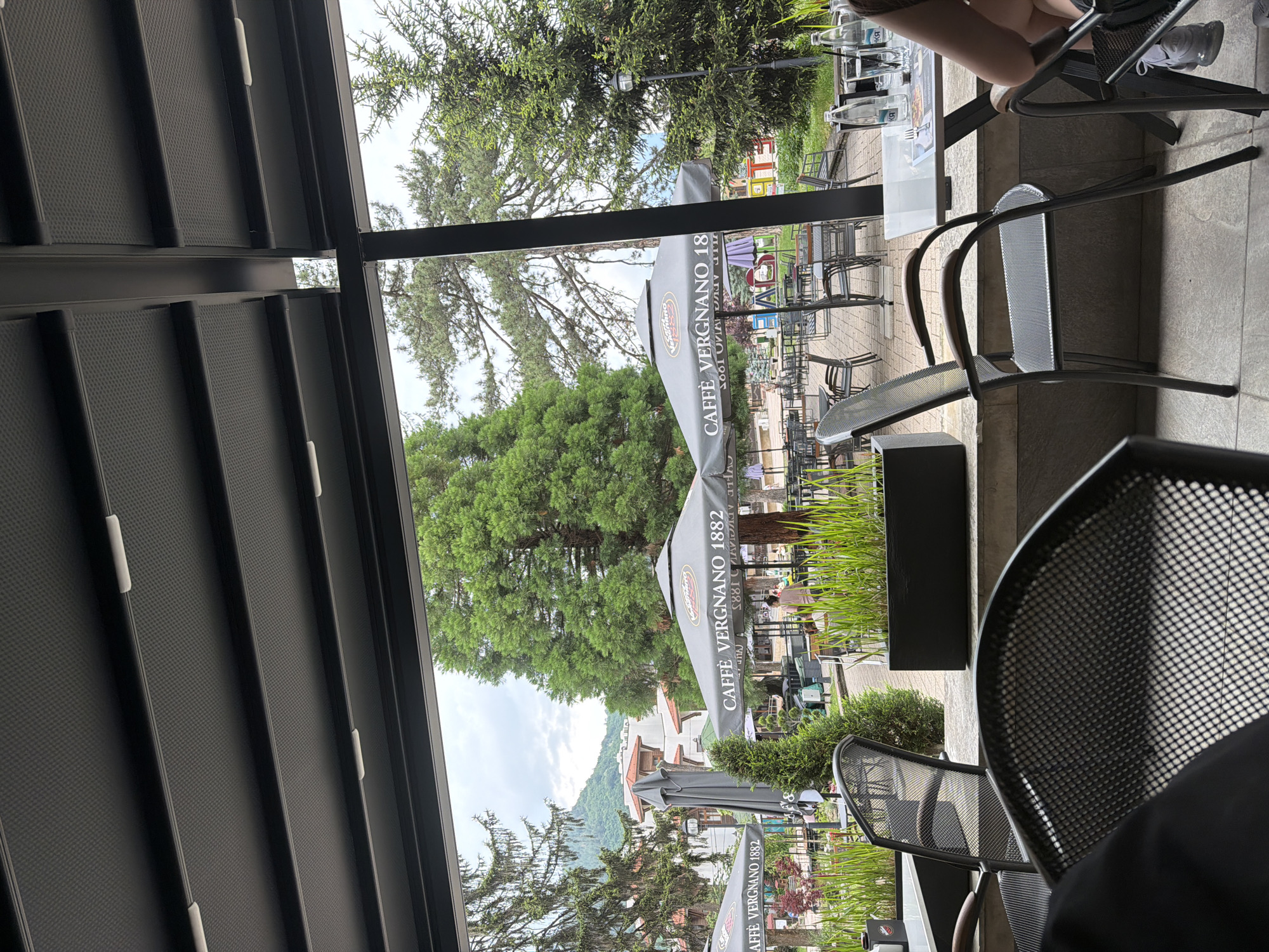 The image depicts an outdoor café patio in Etropole, Bulgaria, viewed from beneath a dark gray, segmented retractable canopy with integrated white lights. In the immediate foreground, dark metal mesh chairs are partially visible, with a person seated at a white table on the right. This table holds two glass bottles of water, two empty glasses, and a menu. Planter boxes filled with tall green grass-like plants line the edge of the patio.
Beyond the railing, the midground reveals a paved outdoor seating area furnished with numerous dark metal tables and chairs. Several large, dark gray umbrellas are prominent, each bearing the white text "CAFFÈ VERGNANO 1882". One individual stands near an umbrella in the center-left, facing away from the viewer.
The background features a diverse landscape. To the left, several tall evergreen trees frame buildings with white facades and red-tiled roofs. A distinct large, densely foliated green conifer tree stands centrally. Further in the distance, a mountain range is visible under an overcast sky. To the right, more evergreen trees and a black streetlamp are present, with colorful block letters partially visible on the ground that appear to spell "HAOS".