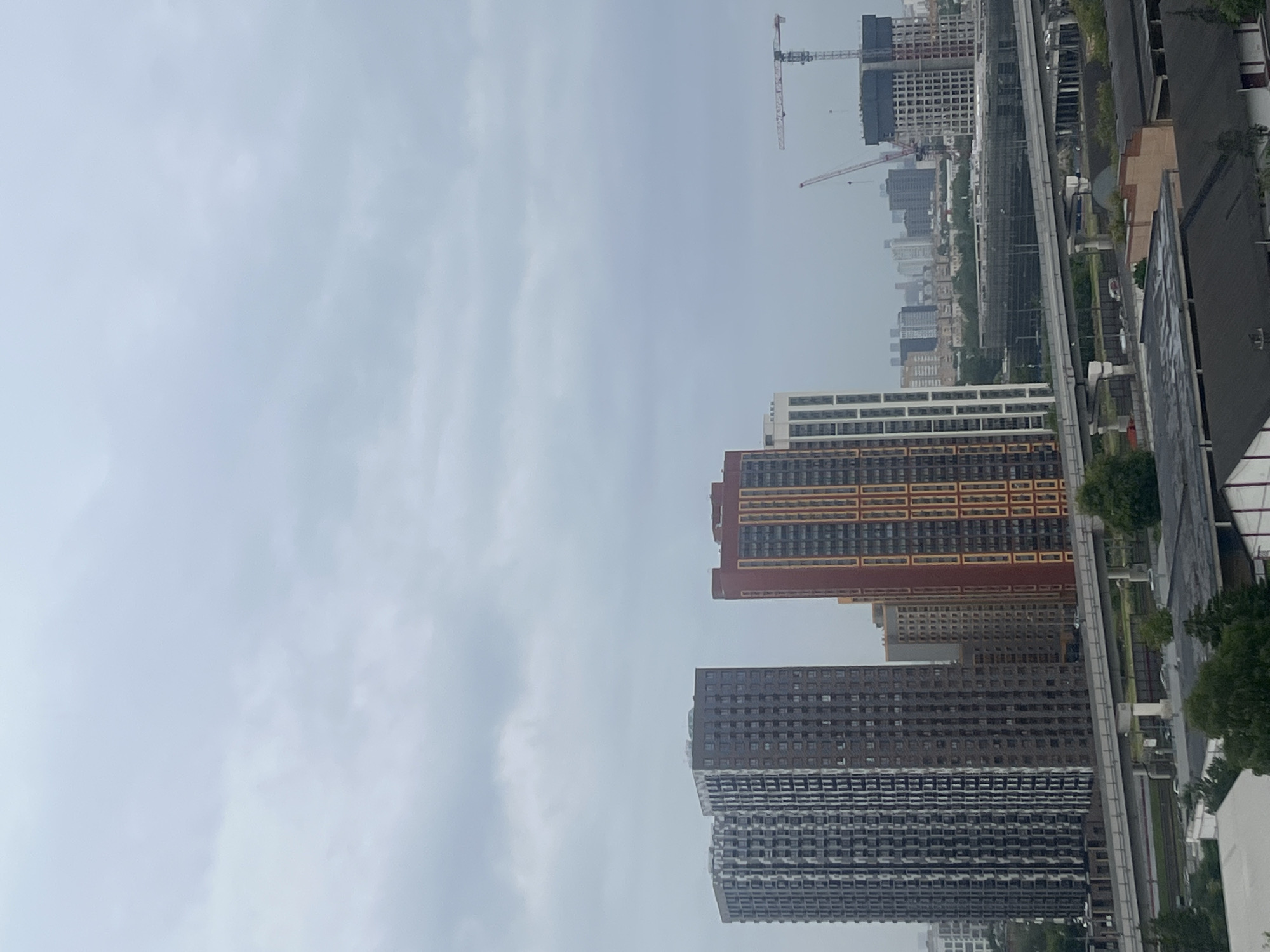 The image depicts a view of the city of Mytishchi, Russia, on an overcast day. In the foreground, several modern high-rise buildings are visible. The tallest are two dark gray buildings with patterned facades, standing side-by-side. To their right is a reddish-orange building with vertical orange and yellow stripes. Further to the right, there are other buildings of varying heights and colors, including a white building with dark vertical accents.
In the middle ground, a multi-level elevated roadway or railway line crosses the scene. Beneath it, there are trees and some lower-lying structures. In the background, a more distant cityscape of buildings is visible, along with two construction cranes. One crane, predominantly pink and white, is large and prominent, while another, smaller red crane is partially visible. The sky is uniformly gray and cloudy, suggesting a dull or potentially rainy day. There are no people visible in the image. The overall impression is of a developing urban area with ongoing construction.