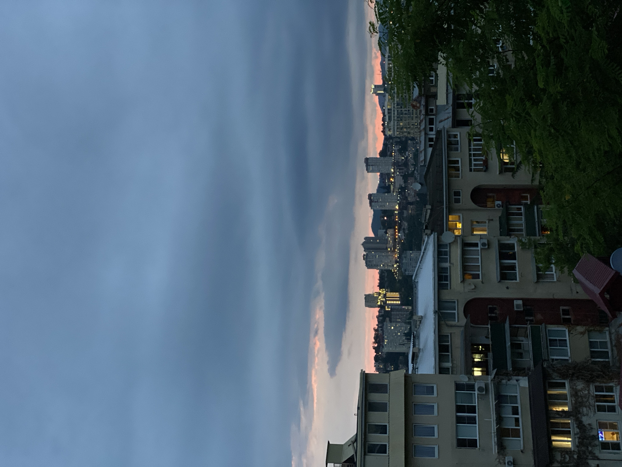 This is a vertical, eye-level, wide shot of a cityscape at dusk, with a focus on residential buildings in the foreground and a more distant view of taller buildings and hills under a cloudy sky. The sky is a gradient of dark blue and gray, with streaks of pink and orange near the horizon, suggesting sunset.
In the foreground, on the left, is a portion of a light-colored, multi-story apartment building with a visible HVAC unit. The windows are dark, but a few show warm interior light. Adjacent to it, and partially obscuring it, is another light-colored apartment building with multiple floors of windows. Some of these windows also emit light. There are air conditioning units mounted on the exterior walls of both buildings. To the right, a cluster of dark green trees partially obscures the view of more apartment buildings. These buildings are also light-colored with visible windows, some of which are illuminated. A dark, sloped roof with a red trim is visible at the bottom right of this section.
In the midground, a densely packed urban area is visible with numerous buildings of varying heights. Several taller, modern buildings stand out, illuminated by internal lights. The cityscape stretches across the horizon, with hills visible in the distance.
The image depicts a residential area within a city, likely during evening hours. The presence of apartment buildings, streetlights, and illuminated windows indicates an inhabited urban environment. The vegetation suggests a temperate climate. The general atmosphere is calm and serene, characteristic of twilight. While the specific location is not visually confirmed in the image, the provided context indicates this is a cityscape in Sochi, Russia. There are no people visible in the image.