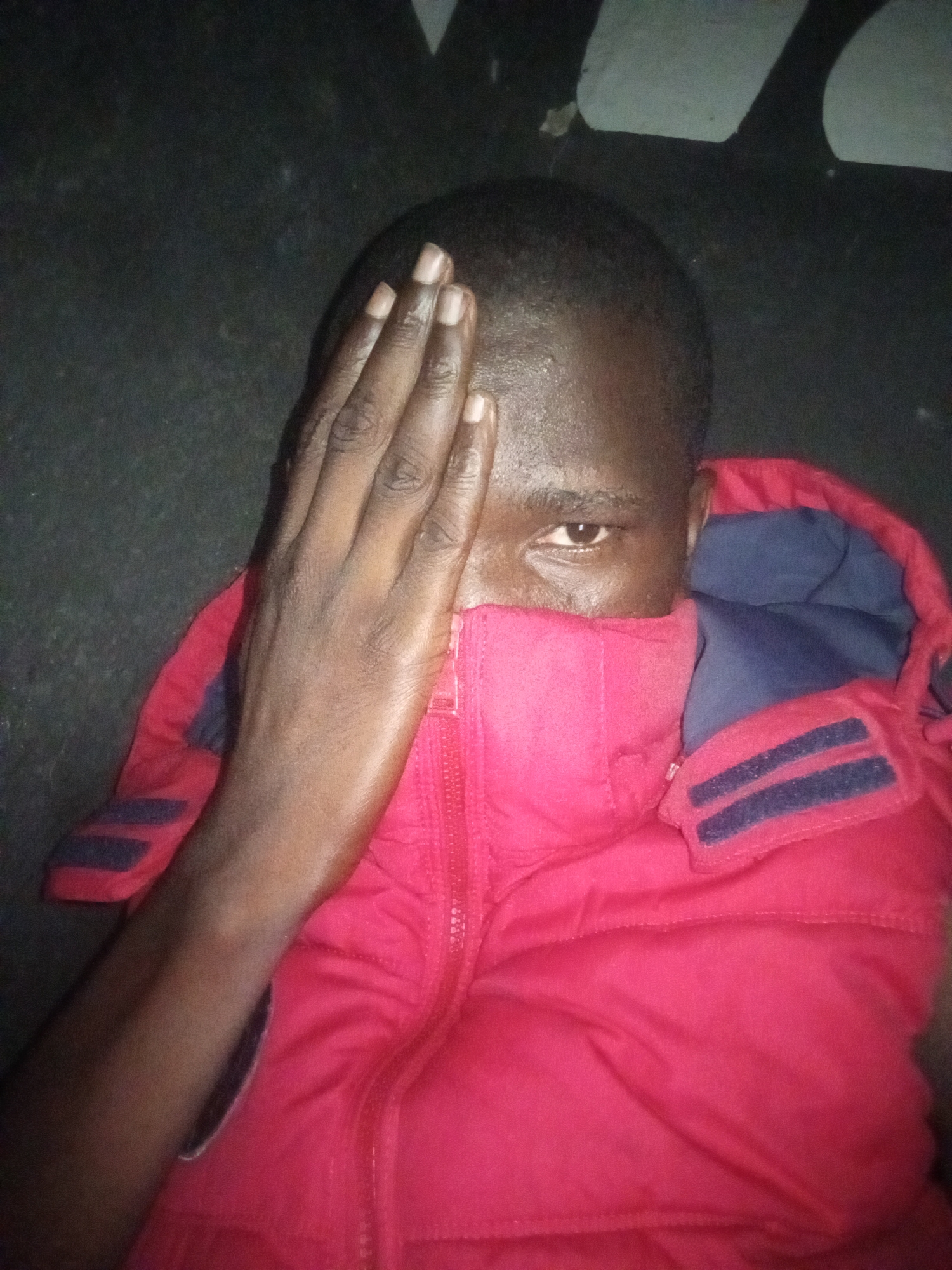 A person is covering half of their face with their right hand. The person has dark skin, short hair, and brown eyes. The person is wearing a red puffer jacket with a blue lining and blue straps on the shoulders. The background is dark. The location is unknown.