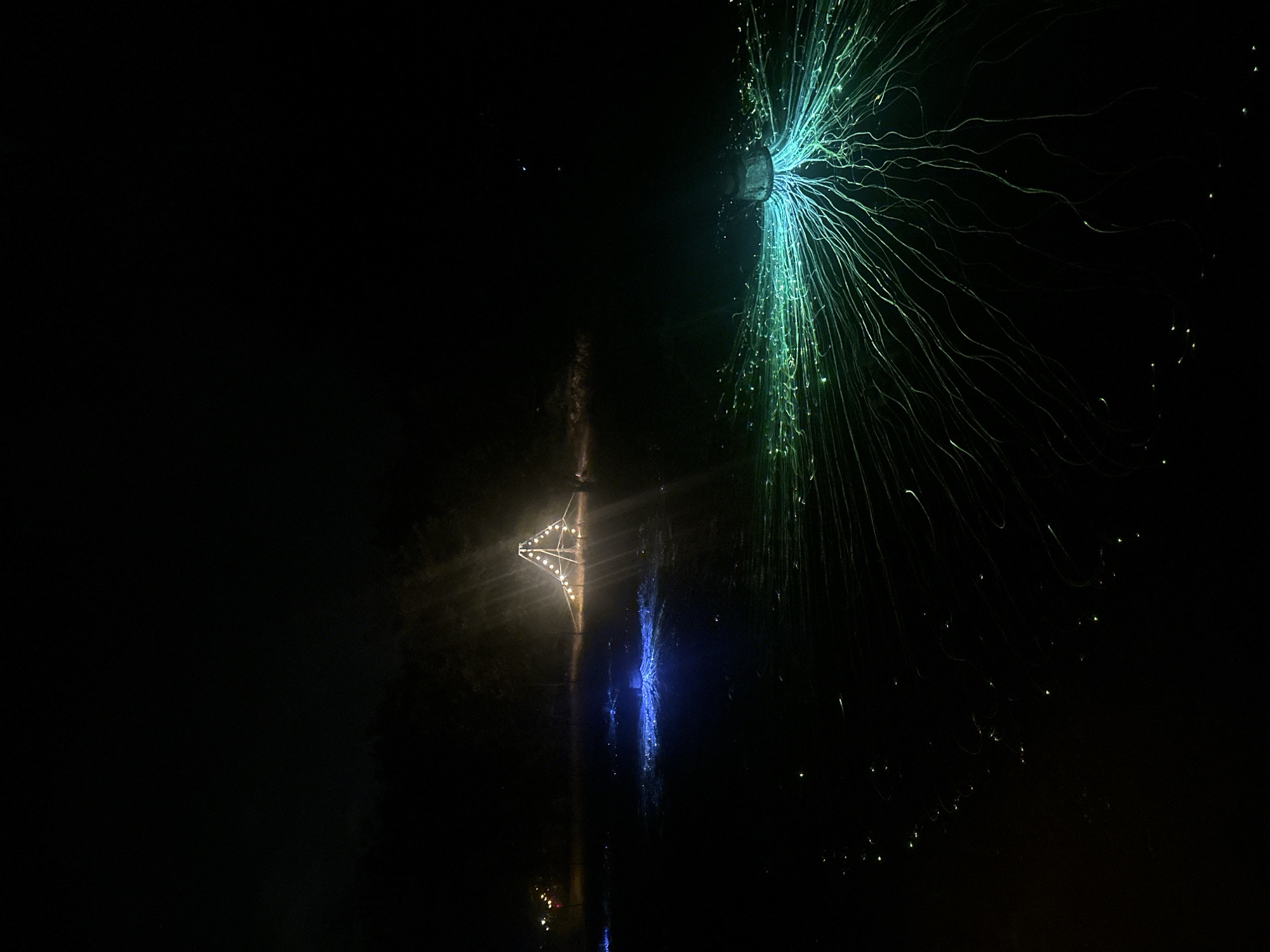 The image depicts a nighttime outdoor scene with several illuminated art installations. In the middle ground, a triangular structure adorned with lights stands prominently. To its left, a patch of ground glows with blue light, and to the right, a larger area features a cluster of green lights from which numerous thin, glowing strands radiate outwards, resembling tentacles or roots. The background is dark and indistinct, with some faint lights visible in the far distance. The overall impression is that of an illuminated exhibit or festival. The location is specified as Bukovik, Montenegro. There are no visible people or animals in the image.