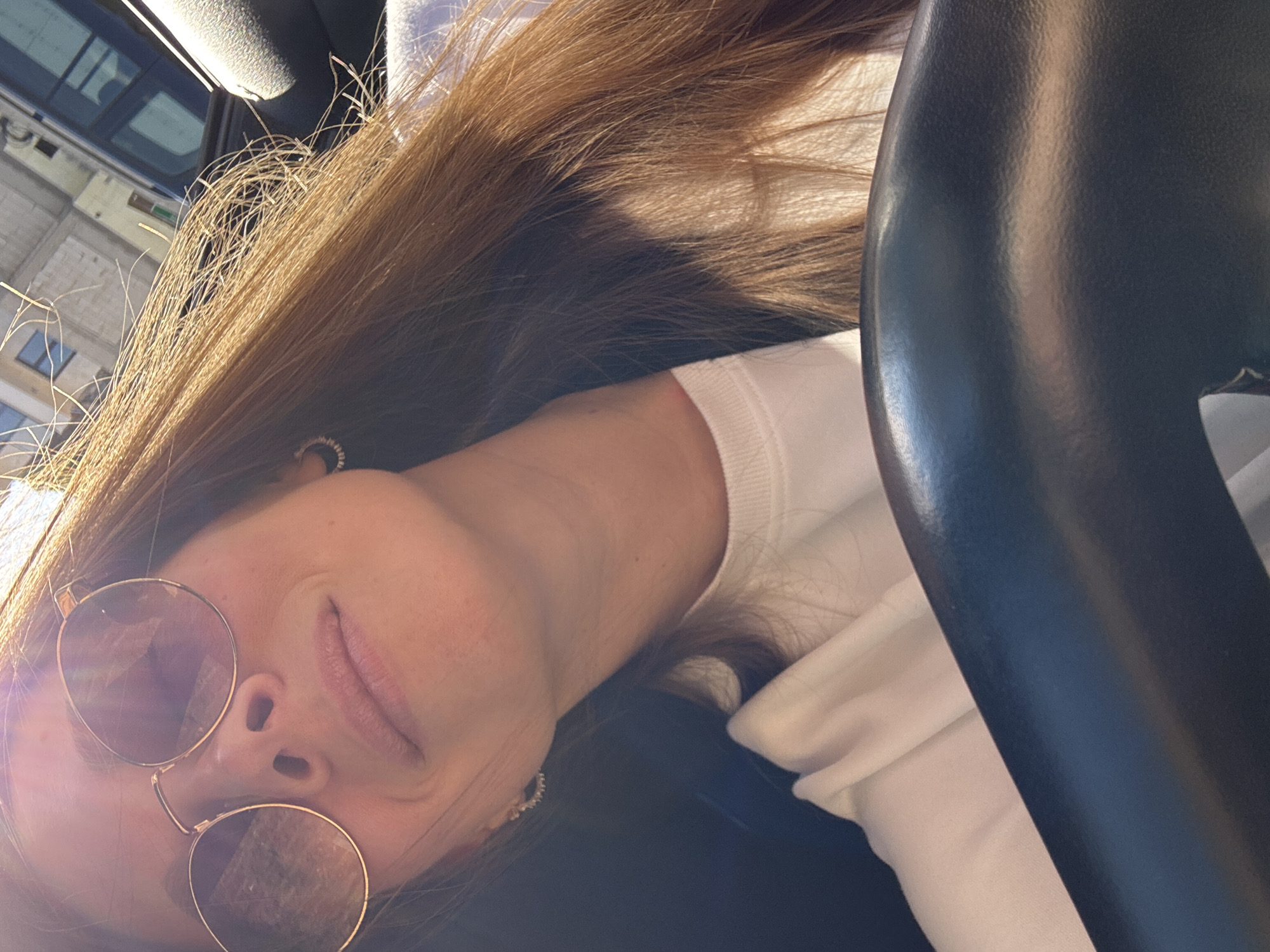 A woman with long, brown hair and fair skin is pictured wearing round, gold-rimmed sunglasses. She is also wearing white clothing. Part of a car's black steering wheel is visible on the right side of the frame. The background visible through the car window suggests an urban environment. While the location cannot be definitively determined from the image, buildings and sky are visible in the reflections of the sunglasses. The city of Budva, Montenegro cannot be confirmed with any certainty.