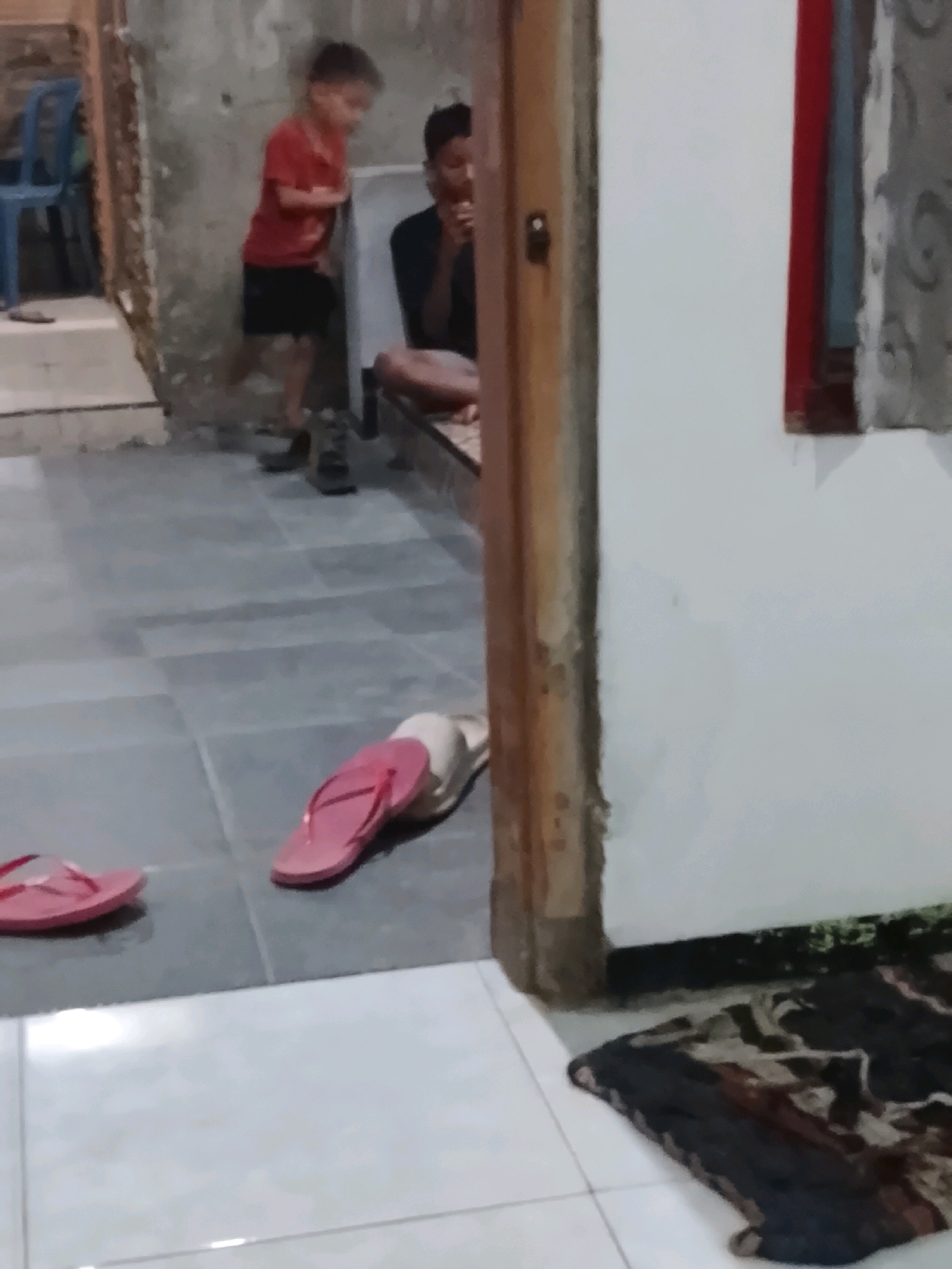 In Ujungrusi, Indonesia, a dimly lit interior scene shows two young boys and a man. One boy, wearing a red shirt and black shorts, stands near a textured gray wall. Another boy, wearing a black shirt, is seated and looking down. A man, also wearing a black shirt, is seated on a mat. The floor is tiled in gray and white. Pink flip-flops are visible on the floor, and a patterned rug lies near the man. A wooden doorframe is to the right of the scene, and a blue chair is visible on the left.
