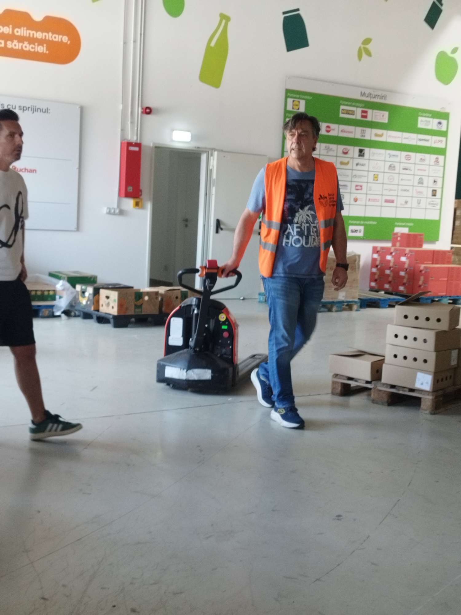 An exterior shot taken at street level in Cernica, Romania, captures a man in an orange safety vest operating a manual pallet jack. The man, with medium-length brown hair, wears blue jeans and a blue t-shirt with a graphic print. He is positioned in the center of the frame, moving a pallet jack with a stack of cardboard boxes on it. To the left of the pallet jack, another man, partially visible from the waist down, is also in the frame, wearing a white t-shirt and black shorts. The background reveals a warehouse-like environment with stacks of cardboard boxes and pallets, suggesting a logistics or distribution center. A large green and white banner displaying logos is visible on the wall behind the man with the pallet jack. The floor is concrete, showing some scuff marks and reflections. The overall lighting indicates daytime, with the scene appearing to be indoors or under a covered structure.