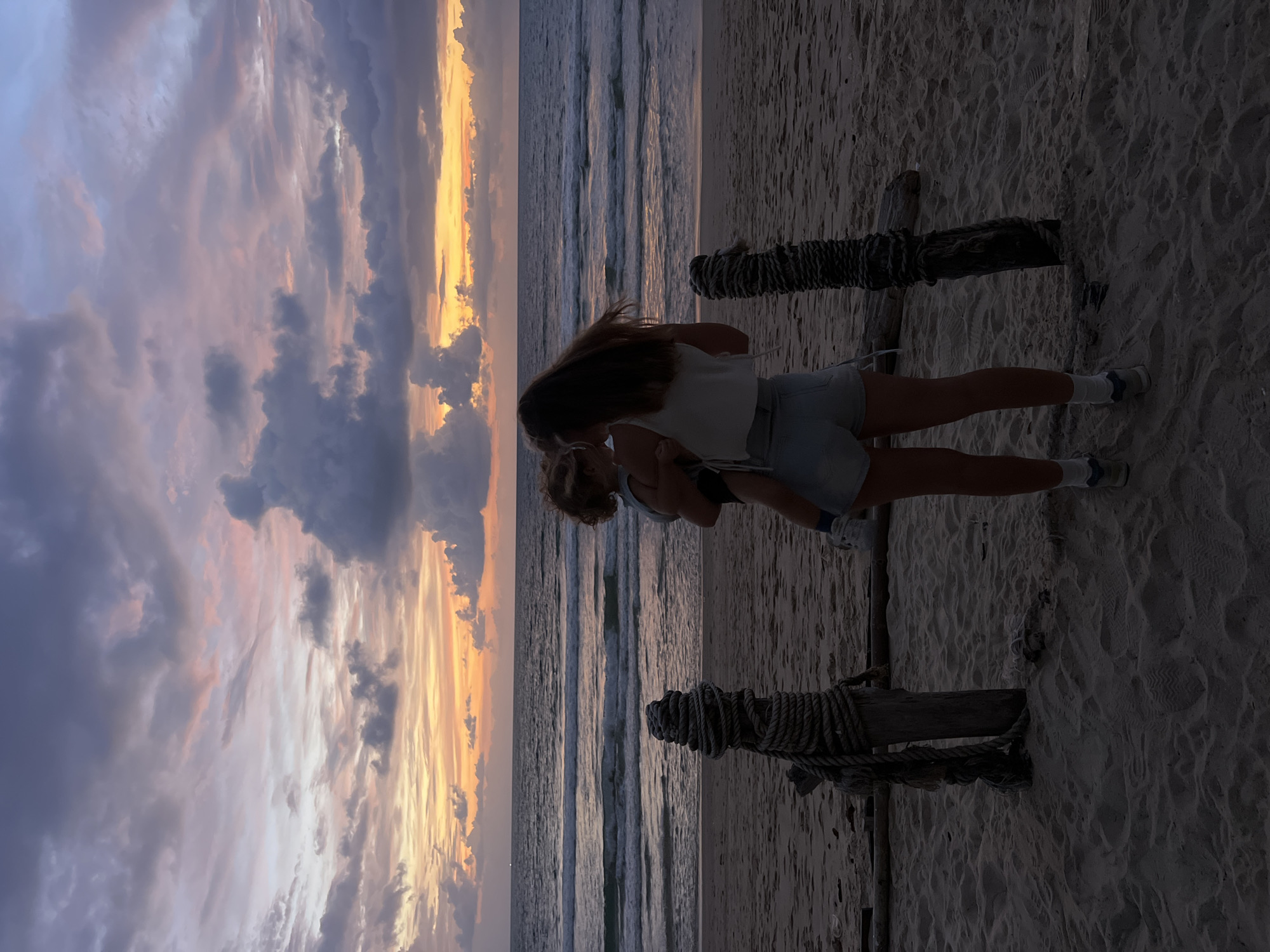 A person holds a child on a sandy beach in Choeng Thale, Thailand, at sunset. The person, facing away from the camera, wears a white top and denim shorts. The child, held in their arms, is partially visible. In the background, waves roll onto the shore under a sky filled with dramatic clouds painted in hues of orange, pink, and purple. Two wooden posts wrapped in rope stand on the beach in the foreground. The scene is silhouetted against the fading light.