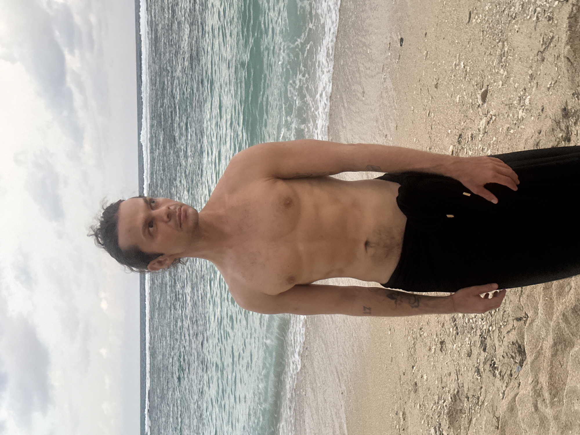 A shirtless man stands on a sandy beach next to the ocean. He is wearing dark pants. The sky is overcast. Based on the prompt information, this photo was taken in Kuta Selatan, Indonesia.
