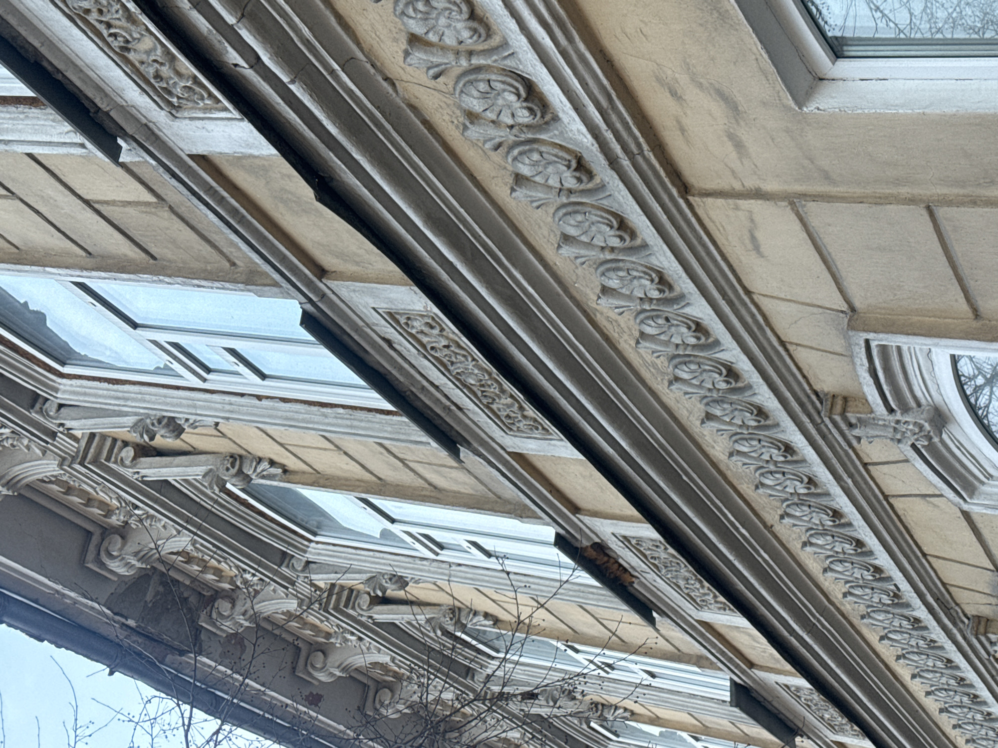 The image depicts the exterior architecture of a building in Krasnoyarsk, Russia. The architecture features decorative trim elements. The image shows the intricate design, with repeating patterns. There are windows, branches from a tree, and elements suggesting some deterioration of the building's facade.