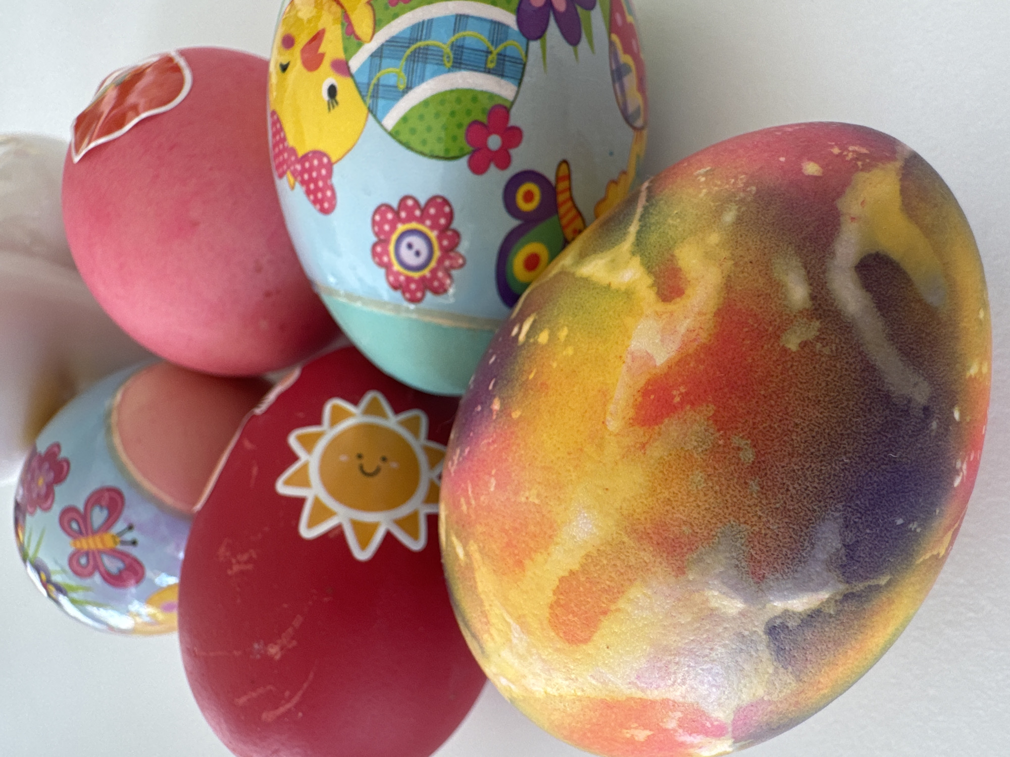 A close-up photograph shows six decorated Easter eggs clustered together against a white background. One egg is pink with a red sticker. Another is light blue with drawings of a yellow horse, flowers, and a butterfly. A red egg has a yellow sun decal. The largest egg has a mottled appearance of yellow, red, and dark gray. The location is unknown and is not confirmed to be in Varna, Bulgaria.
