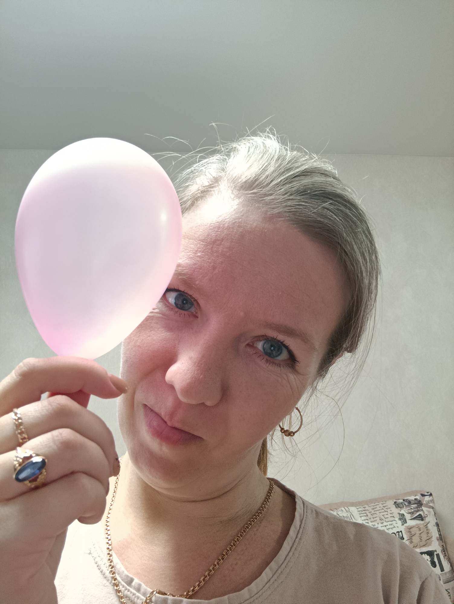 An indoor portrait captures a woman looking directly at the viewer with a slight smile or smirk. She has fair skin, prominent blue eyes, and light-colored, possibly graying hair worn loosely. Her expression appears playful or thoughtful.
She holds a small, translucent light pink balloon in her left hand, positioned to partially cover the left side of her face, near her eye. Her left hand also displays two gold-toned rings: one a simple band and the other featuring a distinct blue oval stone. She wears a casual, light-colored (possibly beige or off-white) top. Further gold-toned accessories include a delicate chain necklace and small hoop earrings with beads.
The background is simple, featuring a plain, light-colored wall and ceiling. Part of a patterned pillow, with indistinct and illegible text, is visible over her right shoulder. The lighting is bright and even, suggesting a daytime setting. The overall scene conveys a casual, personal moment. The location context provided for this image is Chaikovsky, Russia.