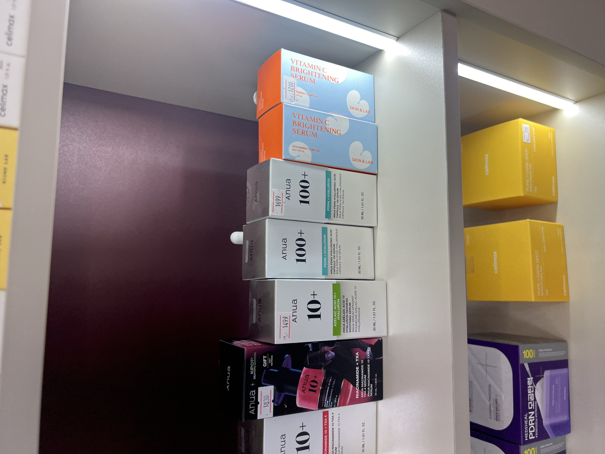 The image shows a retail display shelf in a store. The shelf is stocked with various skincare products, primarily serums and other facial treatments. The products are arranged in their original packaging, mostly boxes.
The top shelf features several Anua brand products, including "Anua 100+" serums with different formulations like "NIACINAMIDE 10 + TXA 4" and "PDRN + HYALURON". One box is a special edition featuring "K-POP DEMON HUNTERS". There are price tags on some of these products, showing prices like 1830, 1499, and 1699. Next to the Anua products are "SKIN & LAB" brand "VITAMIN C BRIGHTENING SERUM" products, with a price tag of 1200.
The lower shelf displays two "MEDIHEAL" brand "PDRN MOISTURE CALMING PAD" products in purple packaging, and two yellow boxes labeled "celimax" with text indicating "PORE + DARK SPOT" treatment. One of these yellow boxes has a price tag of 990.
The lighting in the store is a mix of ambient light and illuminated shelf lighting, casting shadows and highlighting the products. The overall environment appears to be a clean and organized retail space, likely a cosmetics store or a beauty section of a larger department store. There are no people visible in the image. The context of Osh City, Kyrgyzstan, is not discernible from the visual information provided in the image itself.