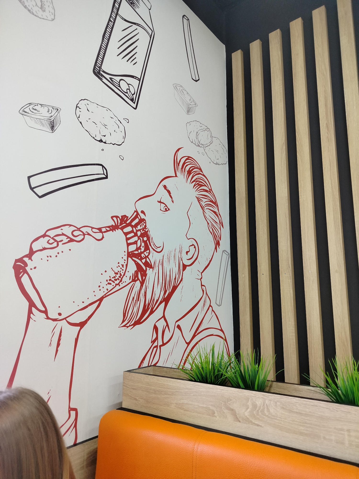 The image shows a wall mural in a restaurant in Taiga, Russia. The mural is a stylized drawing in red ink on a white background, depicting a person with a beard eating a large sandwich. Above the person, various fast food items are drawn, including a carton of drink, a container of sauce, and what appear to be chicken nuggets or similar fried food. To the right of the mural is a decorative wooden slat wall. In the foreground, there is an orange upholstered bench and a wooden planter with artificial grass. The overall impression is of a casual dining establishment.