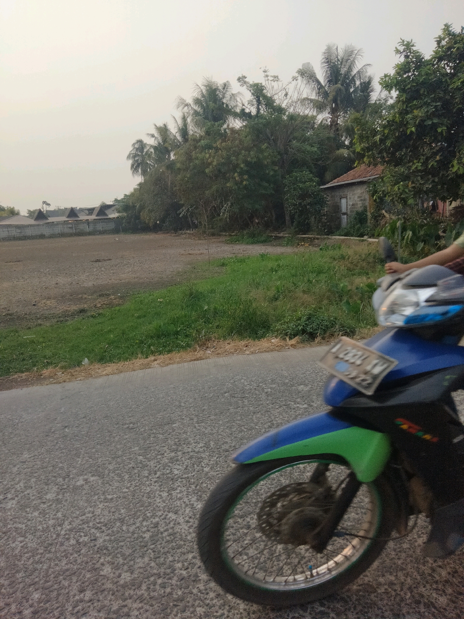 The image captures a roadside scene with a motorcycle in the foreground, obscuring much of the background. The motorcycle is blue and green, with a license plate visible that reads "A 2934 SW" and "09-22". A person's arm and shoulder are visible on the right side of the frame, suggesting they are operating the motorcycle.
The background shows a mostly barren field with some patches of grass, bordered by a concrete fence. Beyond the fence, several buildings with triangular roofs are visible. Further in the distance, a cluster of trees, including palm trees, creates a dense foliage. A small, dilapidated building with a tiled roof and a wooden door is situated among the trees. The sky is overcast and hazy, indicating a potentially polluted or dusty atmosphere. The overall environment appears to be rural or on the outskirts of a populated area. The presence of the motorcycle and the road suggests travel.