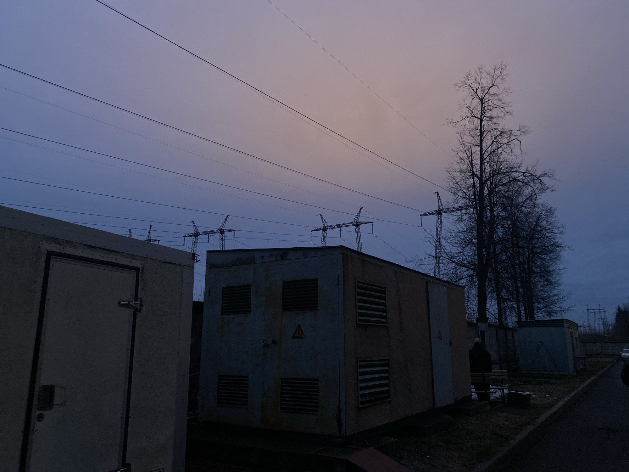 The photo presents an industrial landscape. Several electrical substations stand adjacent to a road. Power lines run overhead, connected to distant pylons. Deciduous trees, devoid of leaves, stand to the right of the substations. The sky is a blend of pale purple and orange. The overall light is dim, suggesting either dusk or dawn. This scene is of a suburban area of Izhevsk, Russia.