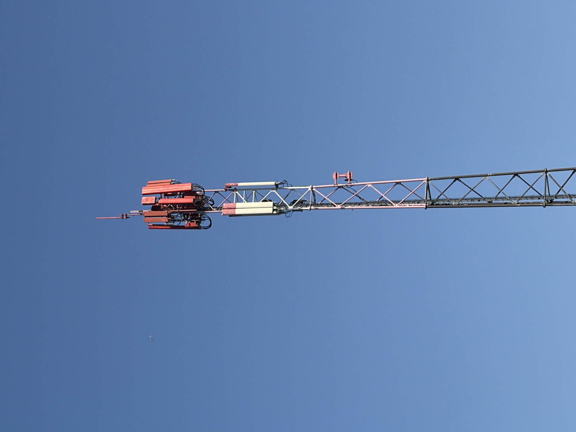 A cellular tower stands tall against a clear blue sky. The tower is a lattice structure with a central pole, painted in shades of gray and pink. Several antennas are mounted at the top of the tower. The top section of the tower features a cluster of red equipment boxes and antennas. Below this, a set of white rectangular antennas are attached. A small, white object, possibly a bird or an aircraft, is visible in the sky to the left of the tower. The bottom portion of the tower shows a criss-cross pattern of metal beams. The overall scene is devoid of any other objects or people.