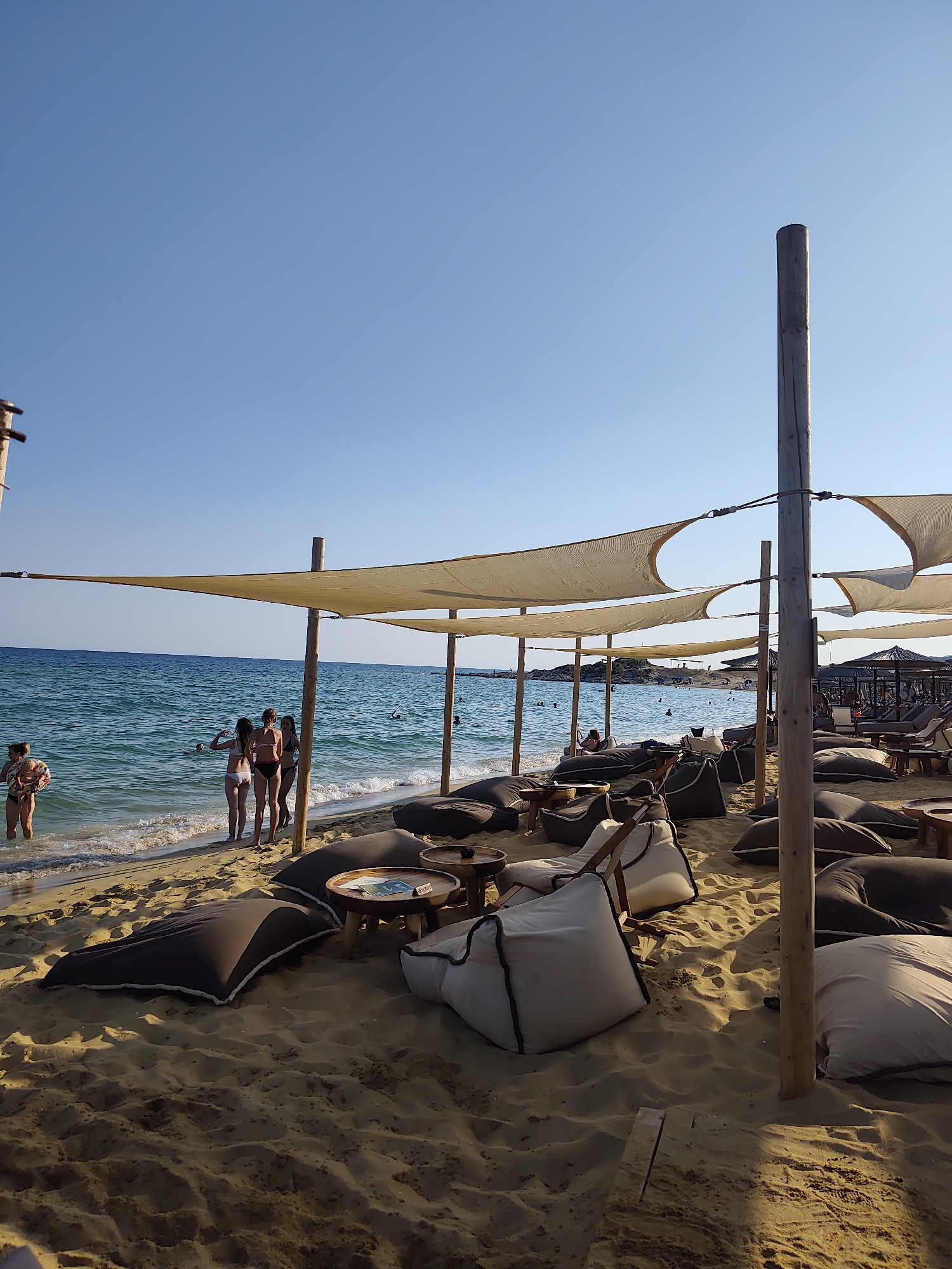 The image depicts a beachside scene in Nea Peramos, Greece, featuring a relaxed, outdoor seating area. In the foreground, numerous large, plush beanbag chairs and low wooden tables are arranged on golden sand. Several people, predominantly women in swimwear, are visible near the shoreline. Some are standing in the shallow water, while others are on the beach. The sea is a vibrant blue-green, with gentle waves lapping the shore. Overhead, canvas canopies supported by wooden poles provide shade over the seating area, suggesting this is part of a beach club or cafe. The sky is a clear, bright blue, indicating a sunny day. In the background, more beachgoers can be seen, as well as some structures, possibly sun loungers or umbrellas, further down the coastline. The overall atmosphere is one of leisure and vacation.