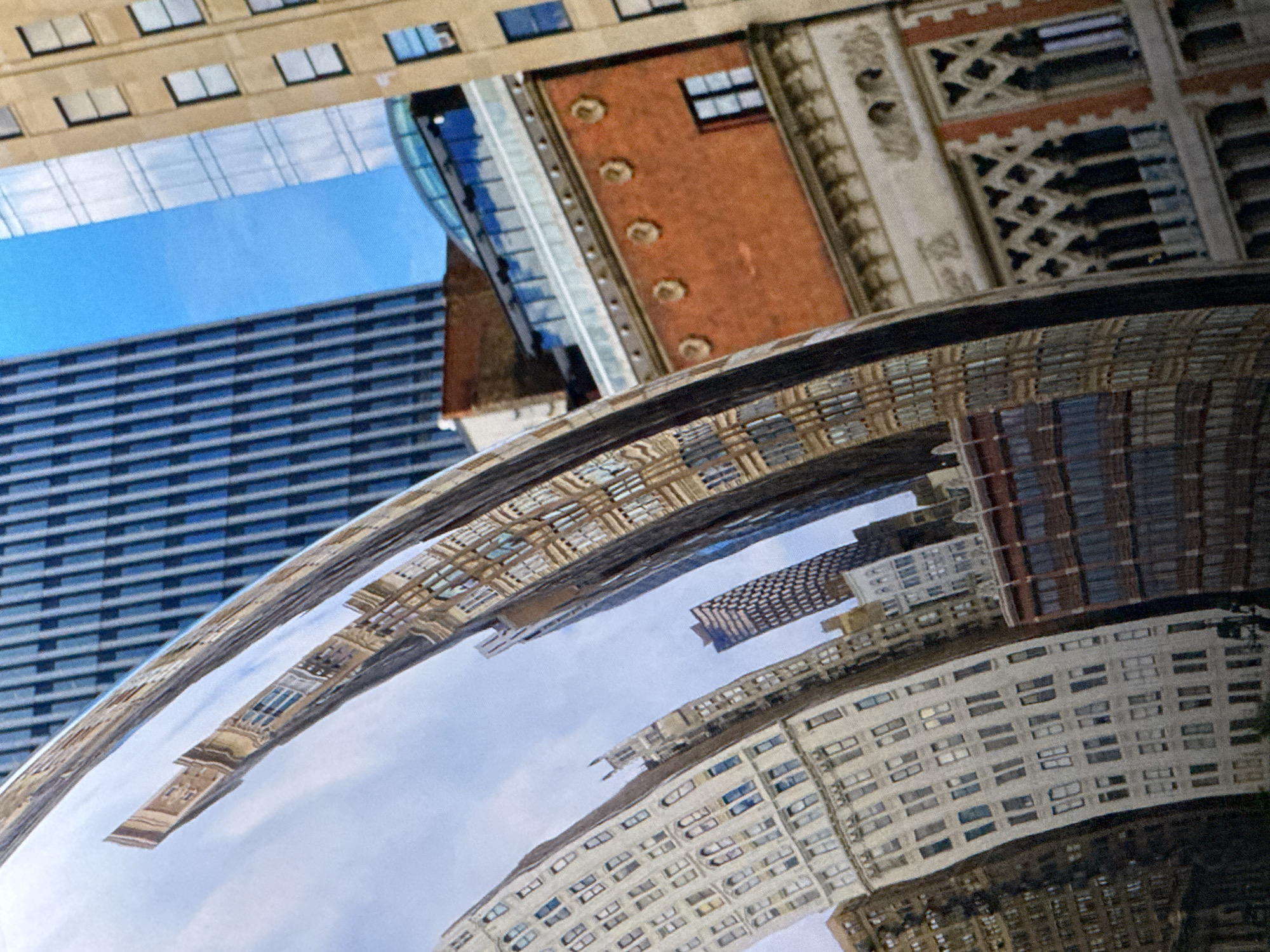 The image is a close-up, distorted reflection of an urban environment captured on a polished, curved metallic surface. The reflection shows a cluster of buildings with varying architectural styles and colors. On the left, a tall, light-colored building with numerous windows is visible, its reflection appearing elongated and curved due to the surface. To the right of this, a darker, narrower building with a patterned facade is reflected. Further to the right, a building with a red brick facade and decorative circular elements is depicted. Above these, parts of taller, modern buildings with glass facades are seen, along with sections of a clear blue sky. The reflection is not geographically located in Saratov, Russia, but rather appears to be a generic urban landscape, possibly taken in a city with a mix of historical and contemporary architecture. There are no people or discernible actions depicted in the image.