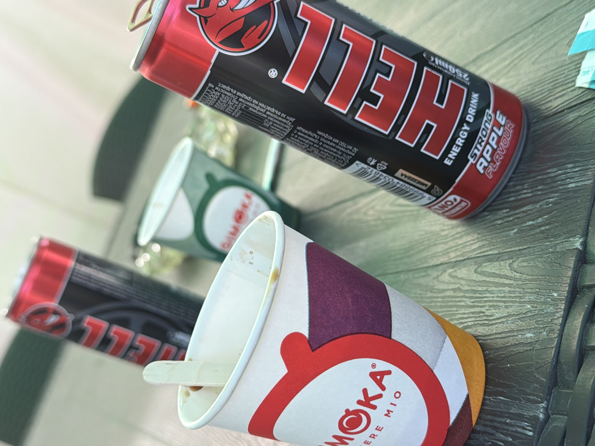 In the city of Razhevo Konare, Bulgaria, a still life photograph captures a table setting with two energy drink cans and a disposable cup. One can, bright red and black, prominently displays the "HELL" logo with a stylized devil mascot, advertising itself as a "STRONG APPLE FLAVOUR ENERGY DRINK." Adjacent to it, another "HELL" energy drink can, partially obscured, is also visible. A white paper cup, branded with "MOKA," sits in the foreground, containing a wooden stirrer and remnants of a beverage. The background is softly blurred, suggesting an indoor or shaded outdoor environment with muted green elements. The overall scene is a casual arrangement of beverage containers, likely captured during a break or refreshment moment.