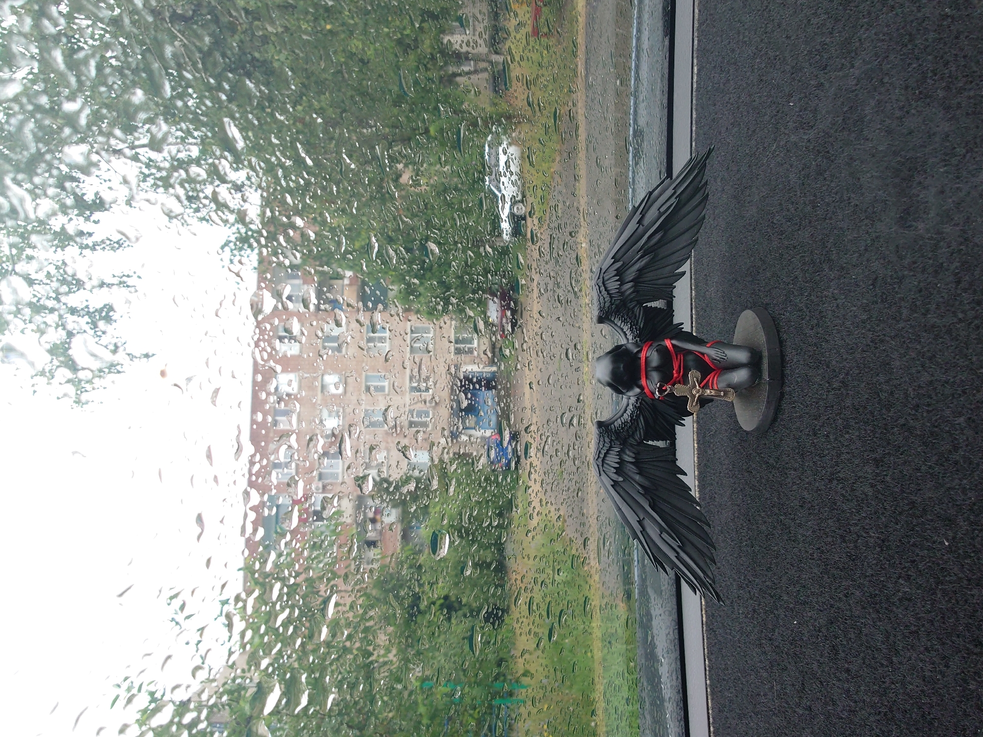A black figurine of a winged figure is positioned on the dashboard of a car, viewed from inside the vehicle. The figurine is kneeling and has large, dark wings. It is adorned with red restraints around its torso and a small cross pendant hangs from its neck. The view outside the car window is obscured by numerous raindrops, creating a blurred effect. Beyond the rain-streaked glass, a multi-story brick apartment building is visible, flanked by lush green trees. Several parked cars are scattered in the foreground, and a patch of grass is also discernible. The overall scene depicts an urban setting, possibly on a rainy day. The city of Sokolohirsk, Ukraine, is not depicted in the image and cannot be inferred from the visual content.