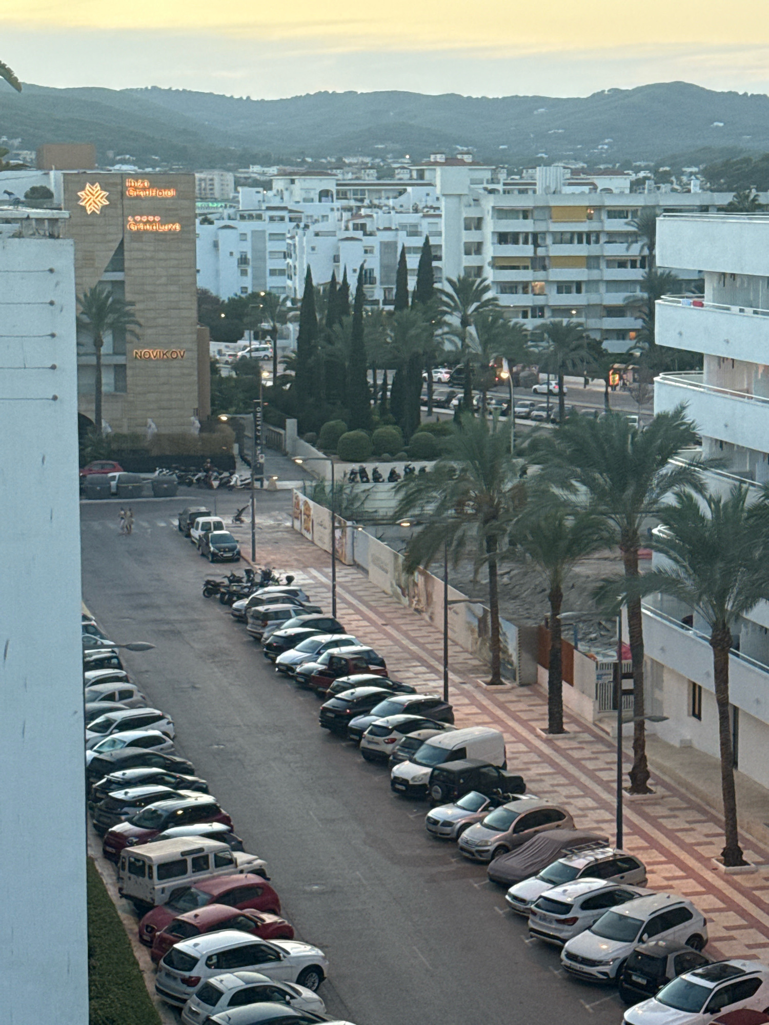 The image depicts a street in Ibiza, Spain, lined with parked cars and palm trees. In the background, buildings of various heights are visible, with a prominent building on the left displaying the "IBIZA GRAND HOTEL" and "NOVIKOV" signage. The sky suggests it is either dawn or dusk. The street is paved with a reddish-brown and white patterned sidewalk. Several individuals are walking on the sidewalk and in the street. The cars are parked in a row along the street, and there are also motorcycles and scooters parked near the larger building. The overall scene is one of a moderately busy urban environment in a Mediterranean location.