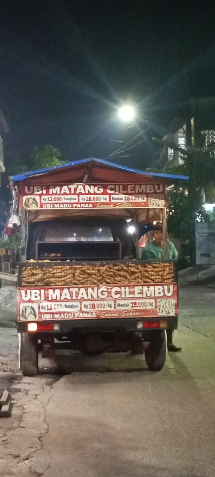 The image depicts a night scene in Setu, Indonesia, focusing on a food truck selling "Ubi Matang Cilembu." The truck is a small, open-air vehicle with a red and blue tarp covering the top, creating a canopy. The back of the truck is loaded with what appears to be sweet potatoes, piled high. A banner on the back of the truck displays pricing information: "Rp 12.000/bungkus" (Rp 12,000 per package), "Rp 28.000/kg" (Rp 28,000 per kilogram), and "Mentah 18.000/kg" (Raw 18,000 per kilogram). Another line on the banner reads "UBI MADU PANAS, ENAAK SEKALI..!" (Hot Honey Sweet Potatoes, Very Delicious!). A young man in a green shirt stands behind the truck, appearing to be the vendor. The truck is parked on a paved road with some cracks, and there are trees and buildings visible in the background, illuminated by artificial lighting. The overall impression is a busy street food vendor operating after dark.