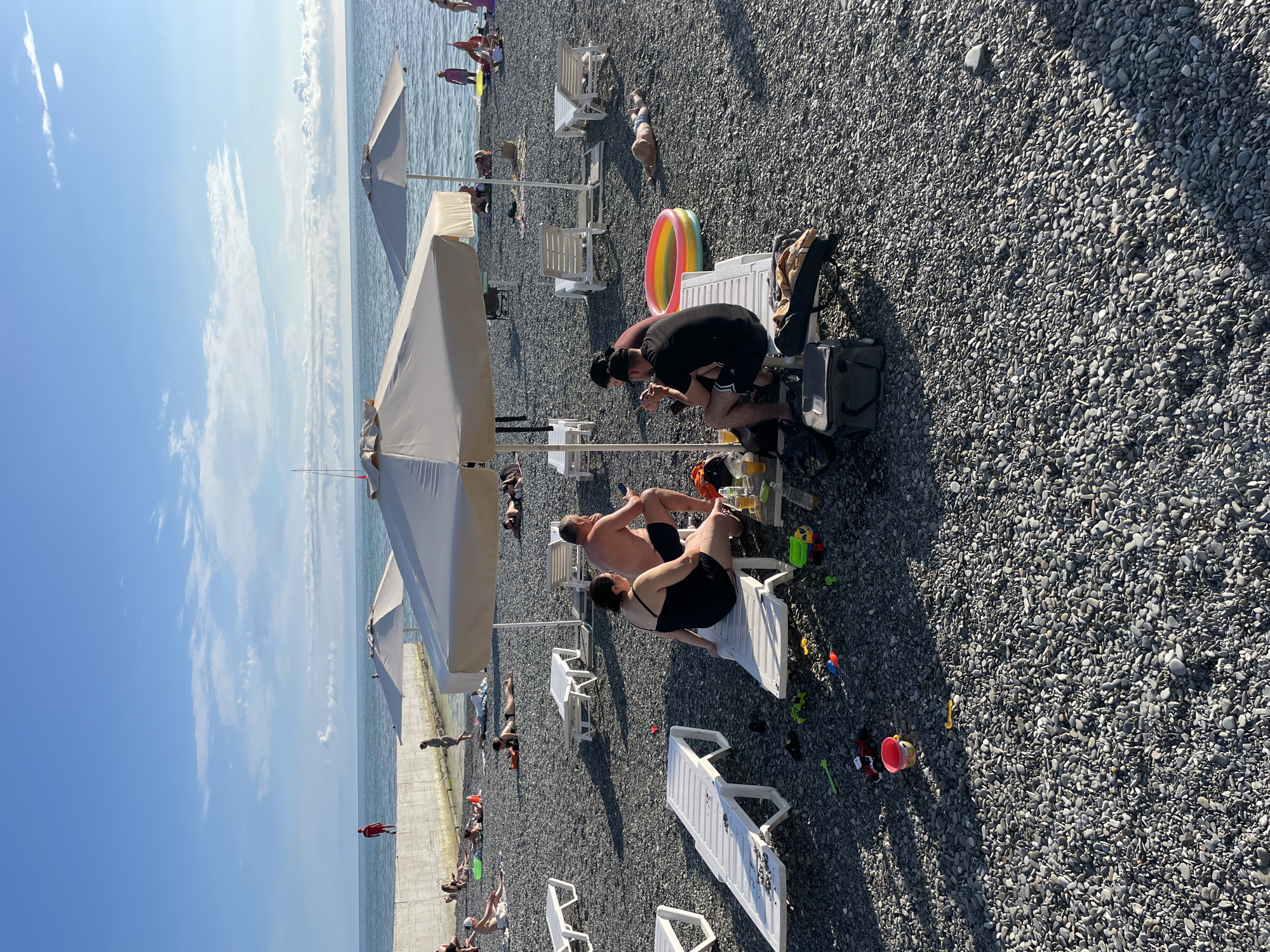 A wide shot of a pebble beach on the Black Sea coast in Sochi, Russia. Several people are sunbathing on white loungers and sitting under large beige umbrellas. In the foreground, a couple is seated on a lounger under an umbrella, with drinks and snacks on a low table. Nearby, a colorful inflatable pool and children's toys are scattered on the pebbles. In the background, more beachgoers are visible, some near the water's edge. A concrete sea wall runs along the coastline. The sky is blue with scattered white clouds.
