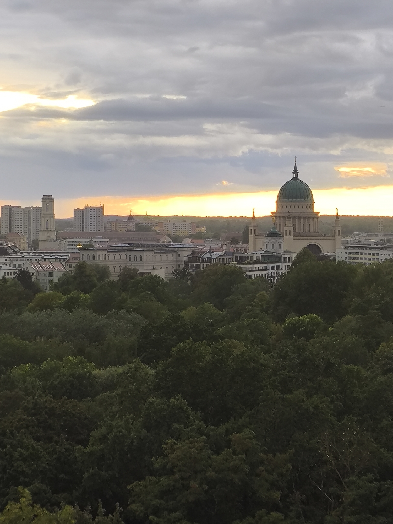 This is a photograph of the cityscape of Potsdam, Germany, taken at dusk. In the foreground, a dense canopy of dark green trees dominates the lower half of the frame, obscuring the ground level. Rising above the trees, a cityscape unfolds. On the left, several tall, modern apartment buildings, appearing light-colored, are visible. To the right of these, a prominent domed building, likely a church or public building with a green dome, stands out. Its classical architecture is evident, with columns and a spire. The background shows a low horizon with more buildings and a range of hills. The sky is overcast with grey clouds, but a sliver of bright orange and yellow light from the setting sun pierces through the clouds near the horizon, casting a warm glow on the upper part of the cityscape and the sky. The overall scene depicts a European city seen from an elevated vantage point, with a blend of modern and classical architecture set against a backdrop of nature and a dramatic sky.