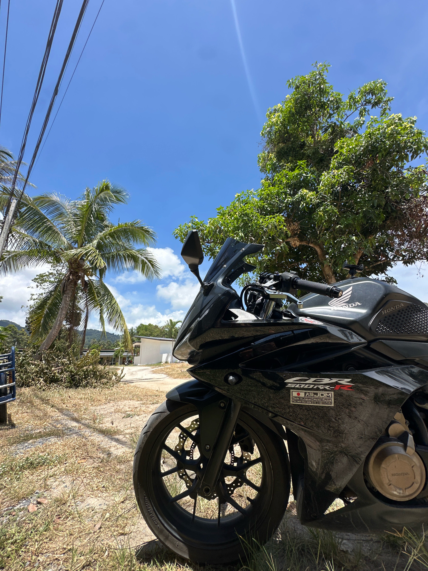 The image depicts a black Honda CBR motorcycle parked on a dirt road in the Ko Pha-ngan District of Thailand. The motorcycle is positioned in the foreground, with its front wheel and part of the fairing visible. Behind the motorcycle, a palm tree leans to the left, and a larger, leafy tree is on the right. A white building with a dark roof is visible in the background, along with some other structures and lush green vegetation, suggesting a tropical or subtropical environment. Power lines are strung across the upper left portion of the frame. The sky is a clear blue with scattered white clouds. The ground is covered with dry grass and dirt. The overall scene suggests a bright, sunny day in a rural or semi-rural area of Ko Pha-ngan.