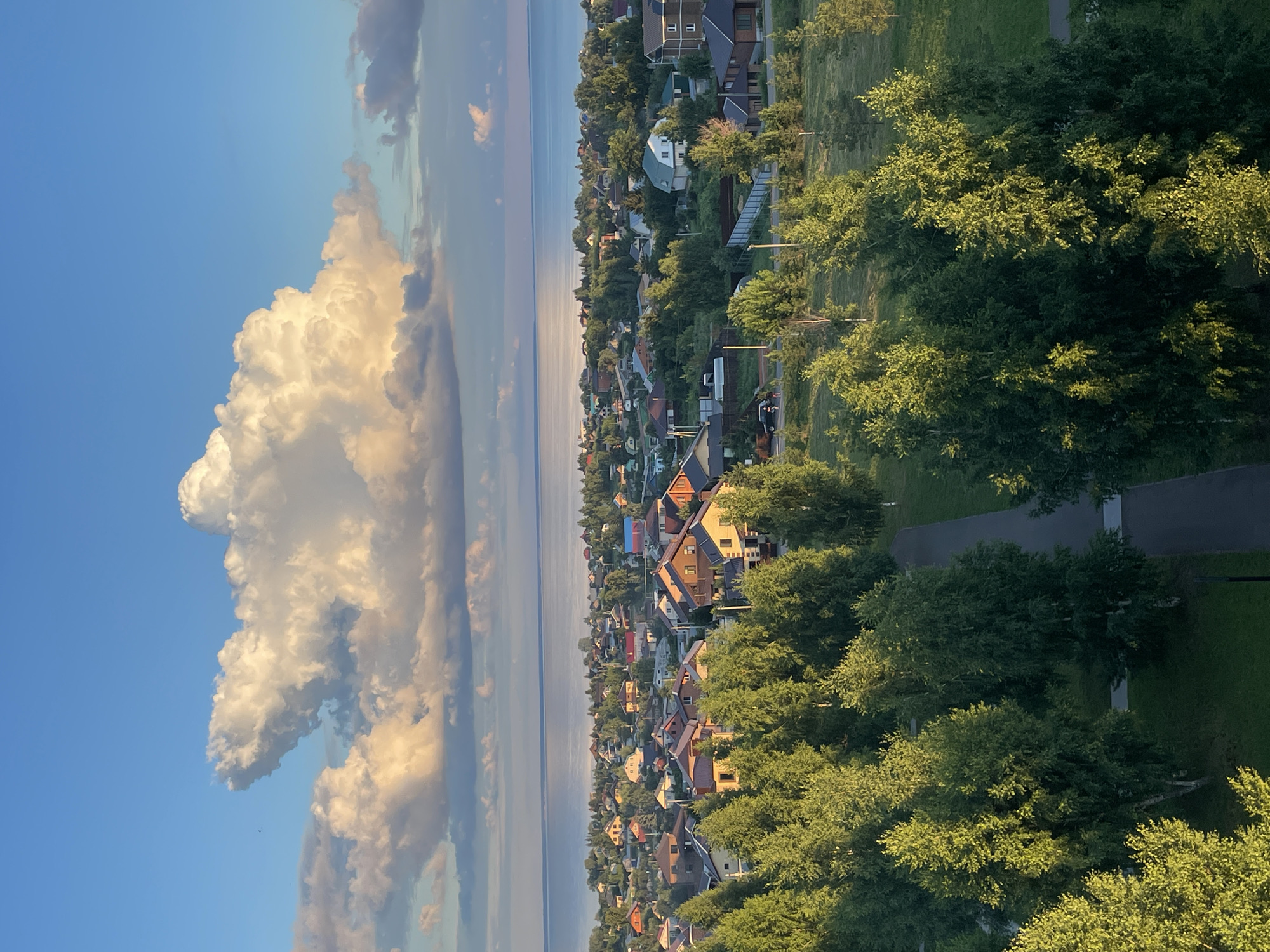 This is an aerial photograph of a suburban area on the coast of a large body of water, likely a lake or sea, under a bright blue sky with a prominent, large cumulus cloud. In the foreground, a well-maintained park with lush green grass and numerous deciduous trees is visible, with a paved walkway winding through it. Beyond the park, a dense neighborhood of residential houses with various roof colors and architectural styles is spread across the landscape, interspersed with more trees. In the distance, the tranquil blue water stretches to the horizon, reflecting the sunlight. The scene appears to be set in Laishevo, Russia, given the architectural style of the houses and the prevalence of birch trees. No people are clearly visible in the image. The overall atmosphere is serene and bathed in the warm light of late afternoon or early morning.