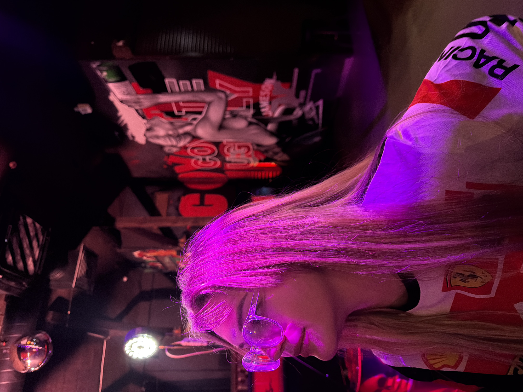 The image captures a person, likely female, with long, straight blonde hair, in what appears to be a bar or club setting in Kazan, Russia. The scene is dimly lit, dominated by dramatic purple and pink artificial lighting that strongly illuminates the person's hair and parts of their face.
The person is wearing a white shirt with red detailing, and the word "RACING" is visible in black text on the shoulder. Another red logo, possibly a shell shape, can be seen on the lower part of their shirt. A unique clear glass or plastic accessory is worn in front of their face, resembling novelty eyewear with two connected bulbous chambers. The person is looking downwards with a faint smile on their lips.
In the dark background, a large banner or poster is prominently displayed. Visible text on the banner includes "JAMESON," and partial words like "GOO GOO" and "LY," alongside red and white graphics and an illustration of a figure. The setting features elements typical of a nightlife venue, including a shiny, reflective disco ball and a bright white LED spotlight, contributing to a vibrant and energetic atmosphere.