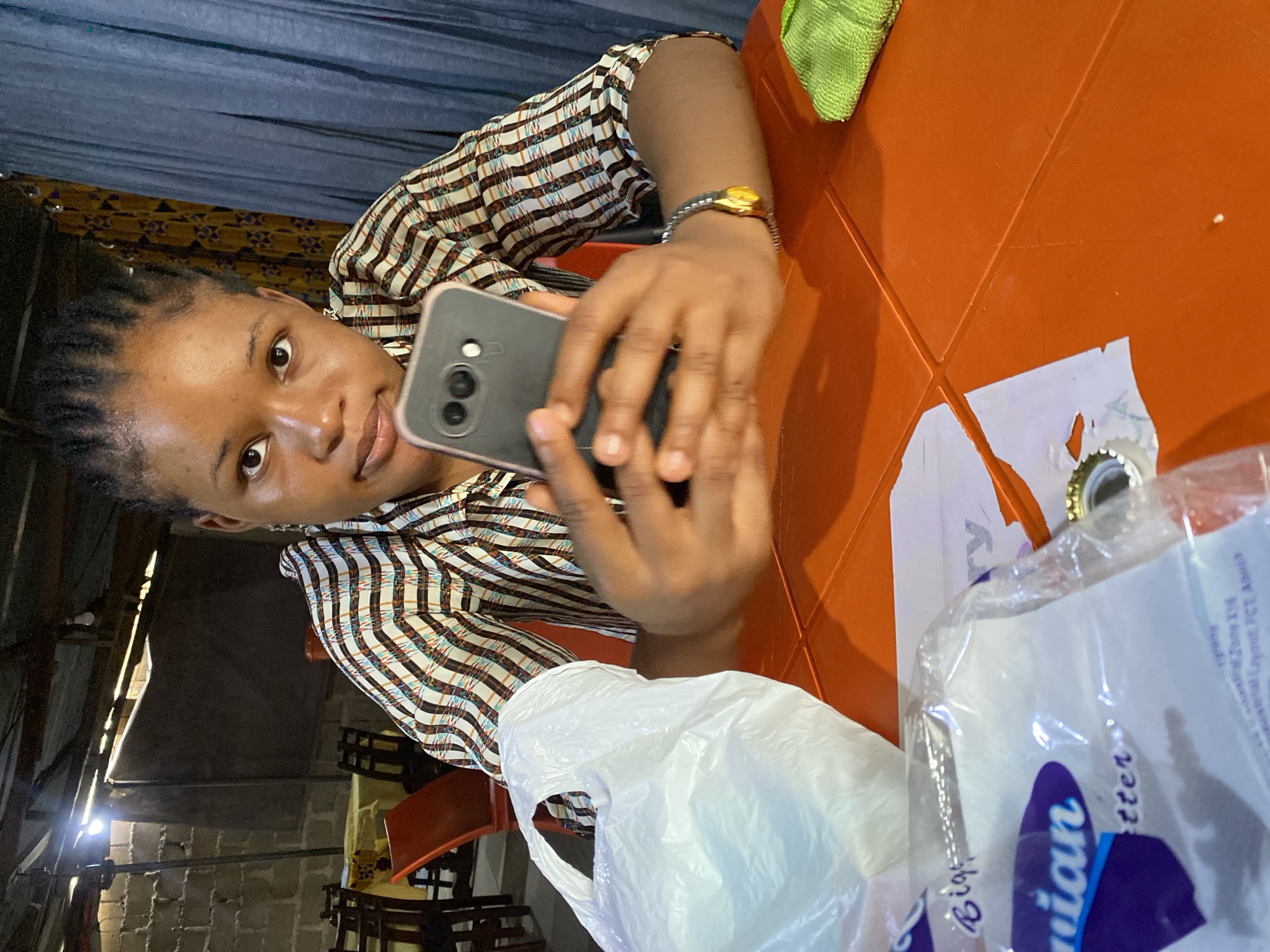 A young Black woman with braided hair sits at an orange table, holding a dark smartphone. She wears a striped collared shirt and a bracelet with a yellow charm. To her left is a white plastic bag, and to her right, a green cloth. The background shows a rough brick wall, wooden chairs, and tables draped with yellow tablecloths, suggesting a casual eatery or outdoor seating area. The lighting indicates it is daytime, possibly late afternoon given the shadows. The overall environment appears to be an informal commercial establishment in Jos, Nigeria, characterized by its utilitarian construction and modest furnishings.