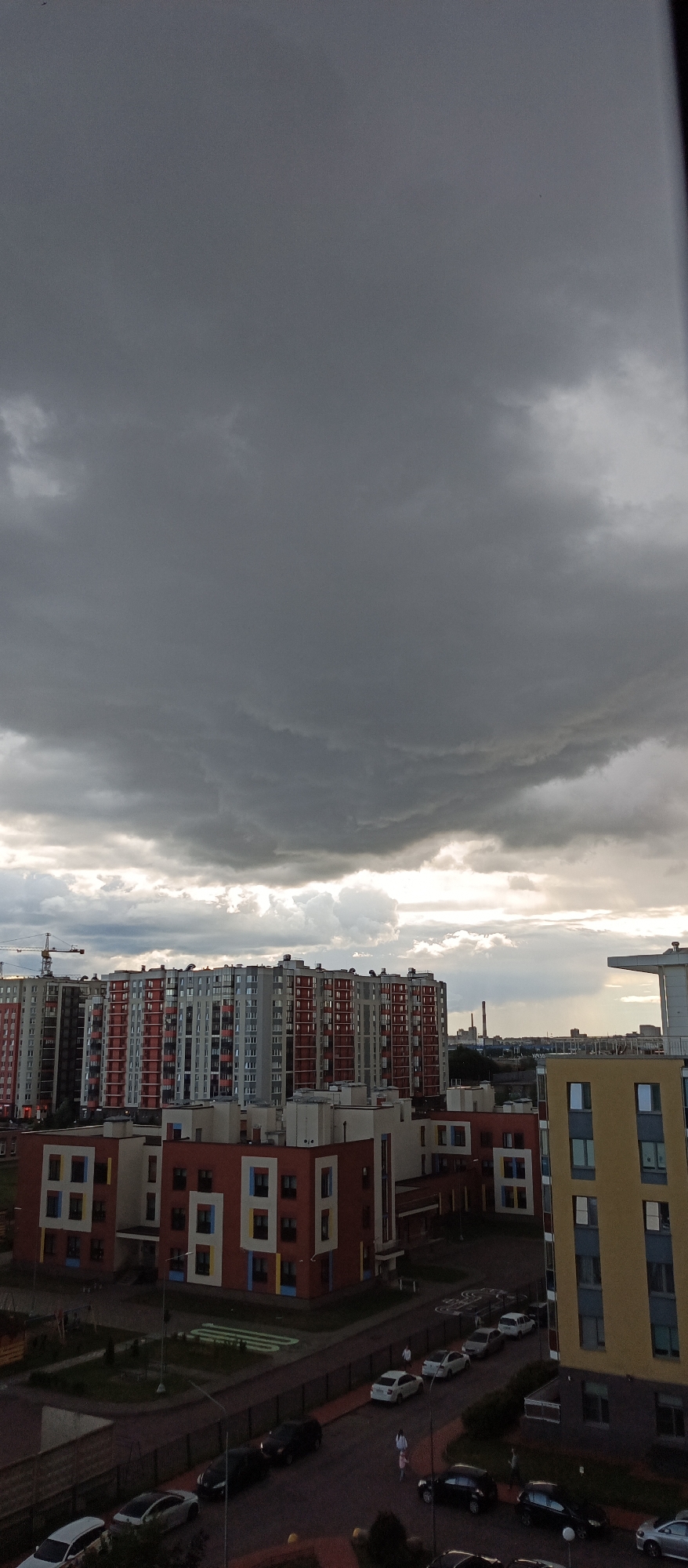 The image depicts an urban landscape in Bugry, Russia, under a dramatic, overcast sky. In the foreground, several modern residential buildings are visible. The closest building on the right is a tall, yellow structure with numerous windows. To its left, a lower building with a red facade and a distinctive pattern of square windows in white, blue, and yellow is prominent. Further back, a row of much larger, taller apartment buildings dominates the mid-ground. These buildings are characterized by their repetitive design, with sections alternating between gray and red or orange facades, punctuated by balconies. A construction crane is visible in the far left of the mid-ground, suggesting ongoing development. The street level shows parked cars, including a white sedan, several dark-colored cars, and a white van. A few individuals are present on the street and sidewalk, including what appear to be two women walking and one person standing. The sky is filled with heavy, dark clouds, with some lighter areas indicating the sun's presence behind them. The overall impression is that of a residential area on a cloudy day.