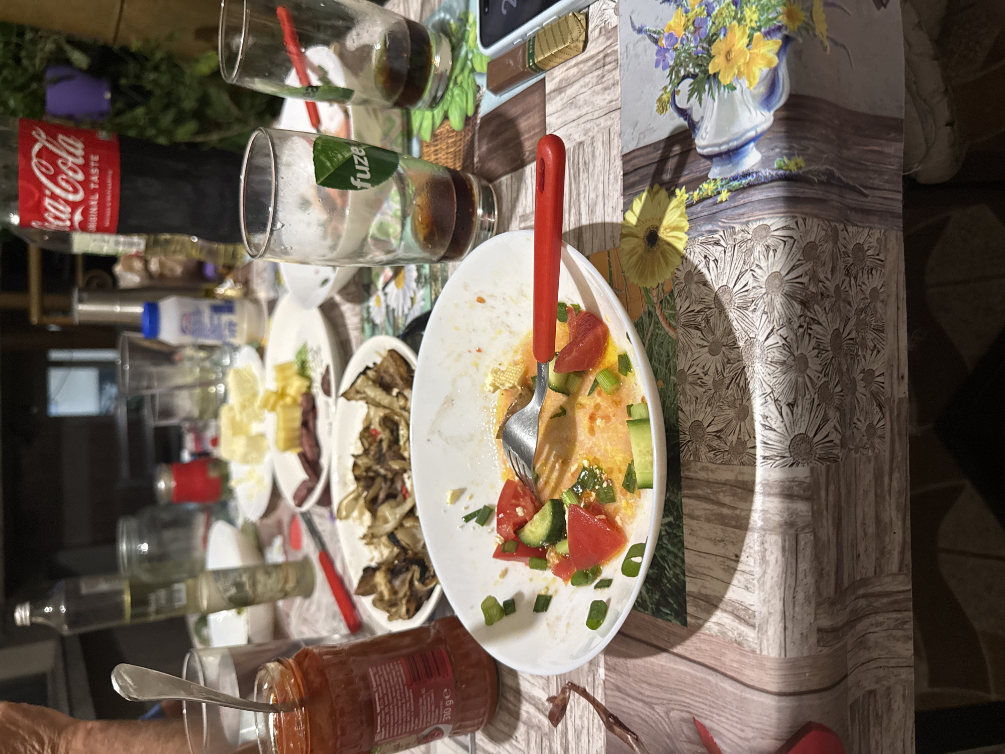 The image shows a dining table laden with the remnants of a meal. There are empty or partially consumed bottles of Coca-Cola and Fuze iced tea. Multiple empty glasses are scattered across the table. Food remains visible on several plates: a plate with fried mushrooms, a plate with cheese, and a white bowl containing remnants of a tomato and cucumber salad with a fork stuck in it. A jar of red sauce is also present. The tablecloth has a printed design with floral elements and a wood-like pattern. Part of a person's hand is visible in the lower-left corner. The location, Sarnevo, Bulgaria, is not ascertainable from the visual information.