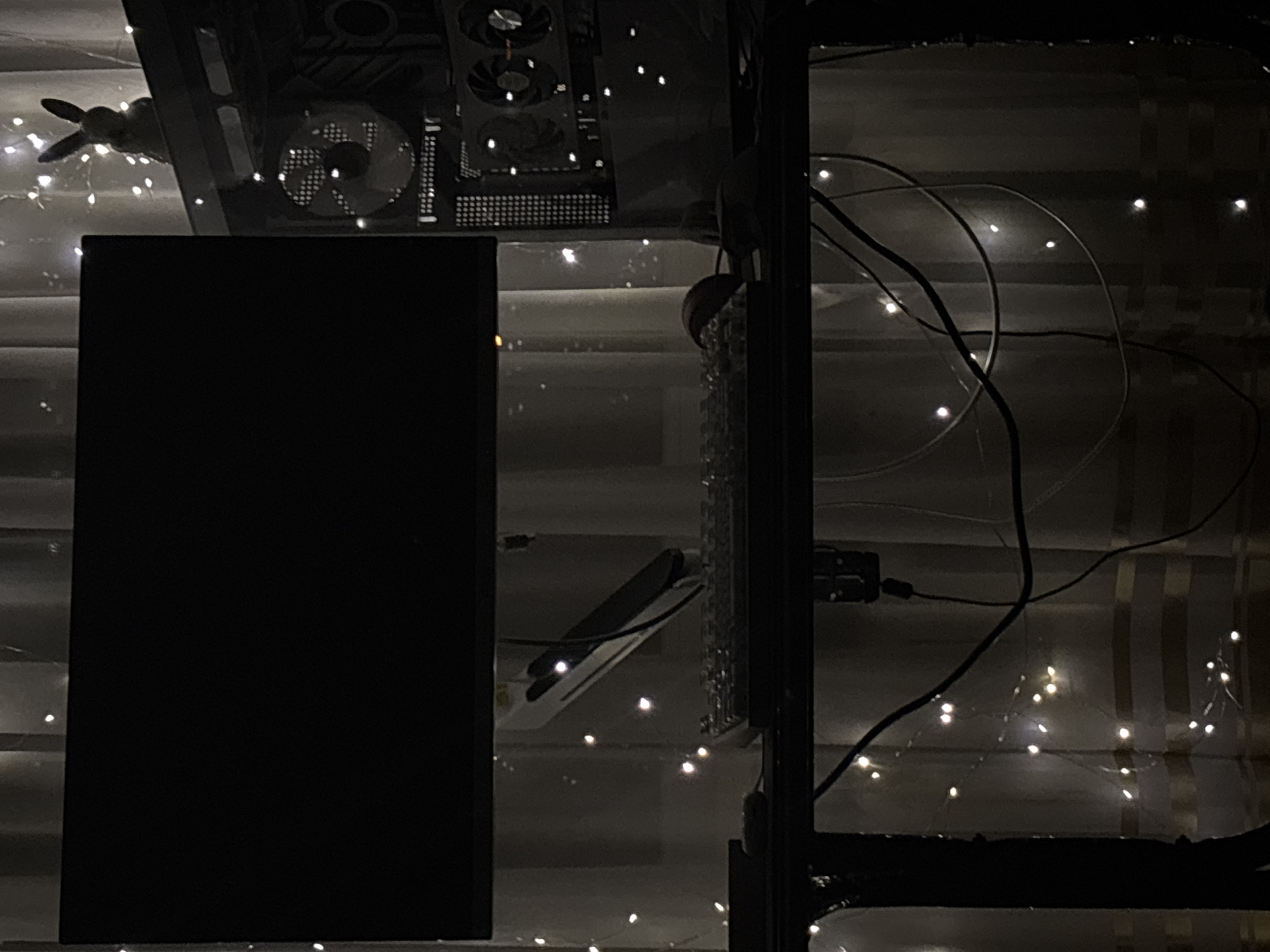 The image shows a dark room with a computer setup. A large monitor is visible on the left, with a keyboard and mouse on the desk below. To the right of the monitor is a computer tower with illuminated fans. A small bunny figurine is perched on top of the computer tower. The background consists of curtains decorated with fairy lights. The overall lighting is dim, with the fairy lights providing a soft glow. The scene appears to be a home office or gaming setup.