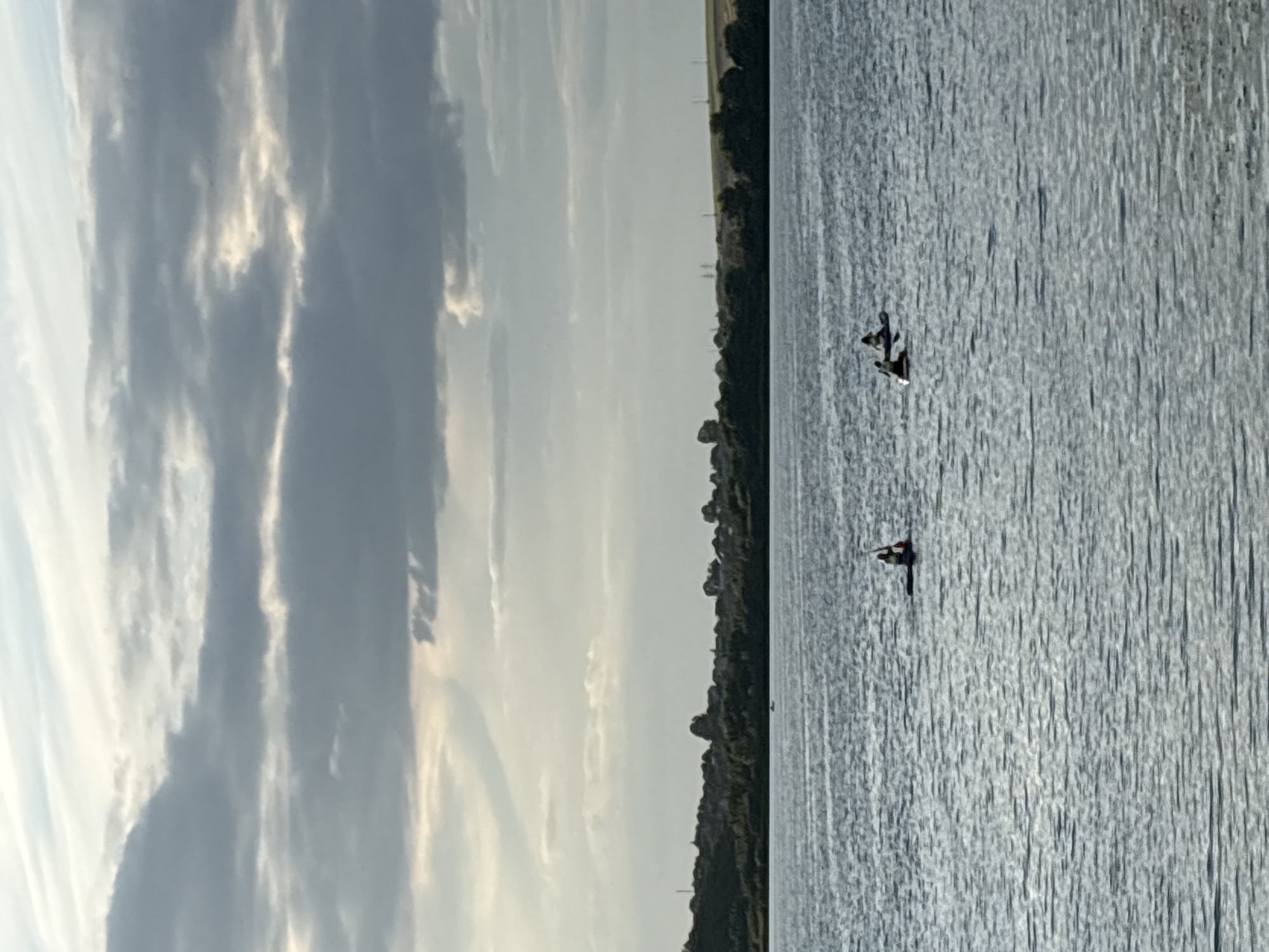The image depicts a wide, calm body of water, likely a lake or reservoir, under a cloudy sky. In the distance, a tree-lined shore is visible, with hills and scattered structures further inland. Three individuals are seen on the water, each on a small watercraft. One person is in a kayak towards the left side of the frame, while two others are further to the right, also in what appear to be kayaks or paddleboards. The water's surface is rippled, reflecting the muted tones of the sky. The scene suggests a peaceful, outdoor recreational setting. Without additional information, the specific location within Ternopil, Ukraine cannot be determined from the image alone.