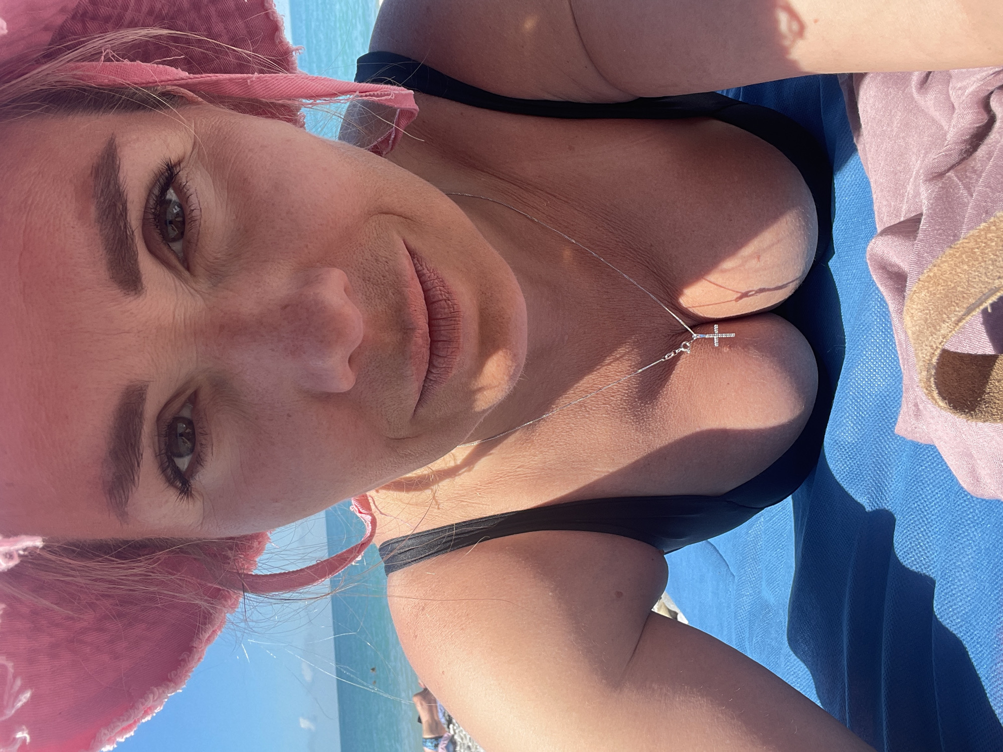 A woman lies on a blue towel on a beach, her head propped up by her arms. She wears a pink bucket hat and a black bikini. A silver cross necklace rests on her chest. The ocean and a hazy shoreline are visible in the background.