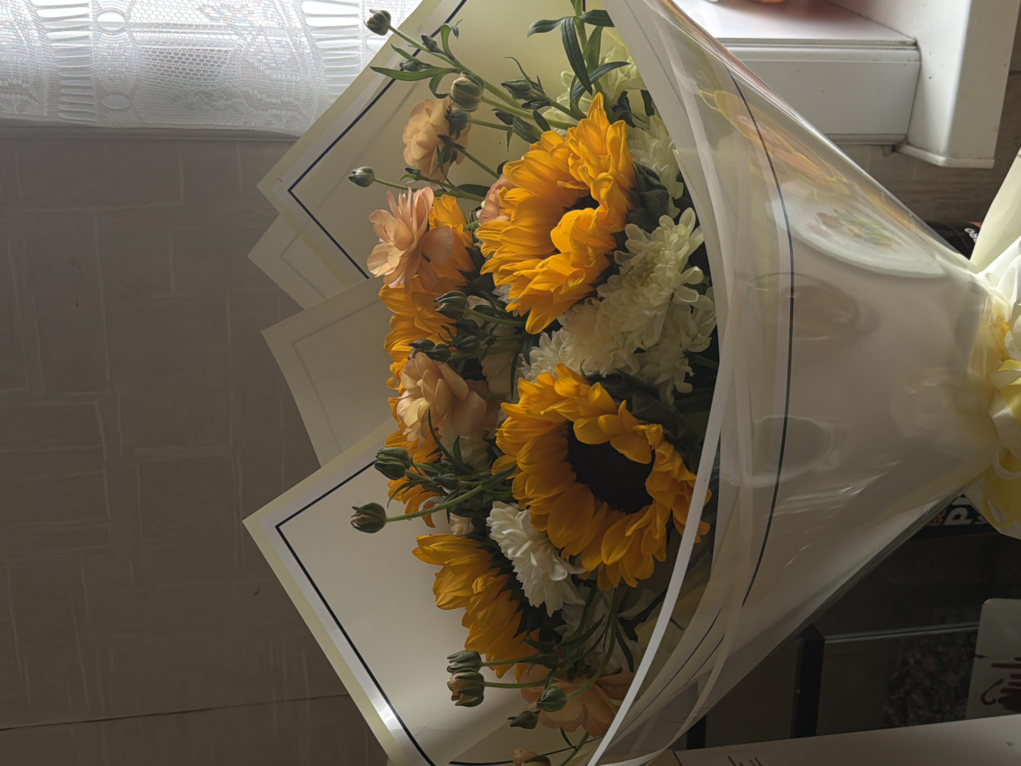 A bouquet of sunflowers and other flowers is wrapped in clear plastic and a light-colored paper liner with a black border. The bouquet is positioned to the right of the frame, with the flowers facing toward the viewer. The background features a textured wall to the left and a window with a white frame and lace curtain at the top of the frame. The location in the city of Perm, Russia, cannot be determined from the image.