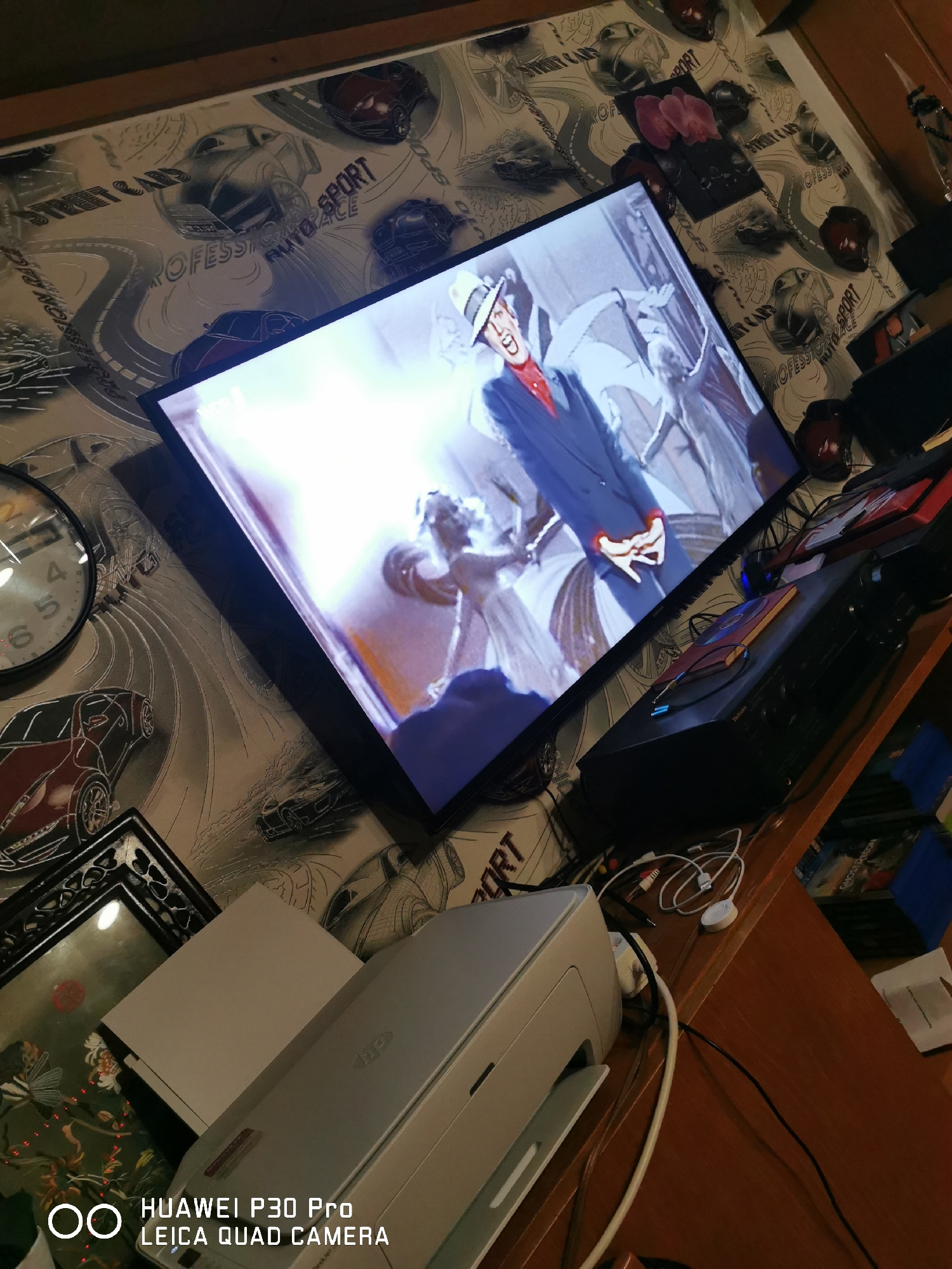 The image depicts an indoor scene, likely a room, with a television screen displaying a film or show. The television is positioned at an angle, angled on what appears to be a wooden desk or shelf. The wall behind the television is covered in wallpaper with a design featuring race cars and tracks. To the left of the television, a round clock with visible numbers is mounted on the wall. In the foreground, a white printer or scanner is visible, with cables connected to it. The overall impression is of a somewhat cluttered living space or entertainment area.