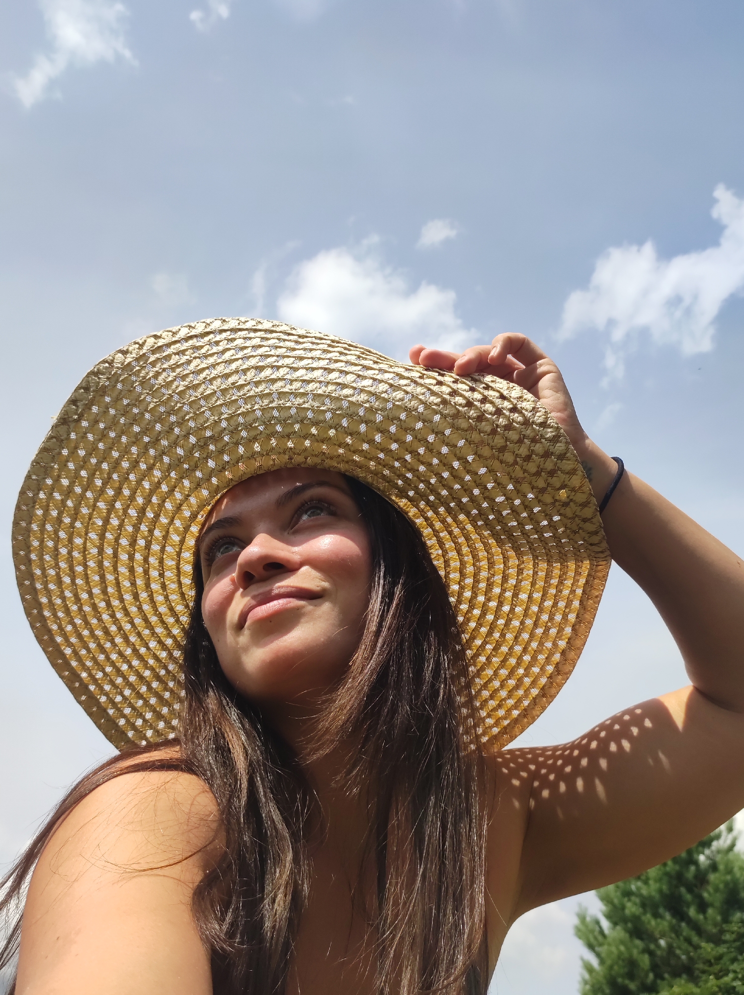 In this low-angle, outdoor, eye-level medium shot, a woman with long, dark brown hair is looking up and to the left, her gaze focused on something beyond the frame. She is wearing a wide-brimmed, woven straw hat that casts dappled shadows on her face and arm. Her left hand is resting on the brim of the hat, and her right arm is bent, with her hand behind her head. The background consists of a bright, blue sky with scattered white clouds. Sunlight illuminates her face, highlighting her features, and creates a warm glow on her skin. A small portion of green foliage, likely trees, is visible in the lower right corner of the frame. The scene appears to be captured on a sunny day, possibly in a park or open area. The woman's expression suggests a moment of contemplation or enjoyment. There is no indication of the city of Фоки, Russia, within the image itself.