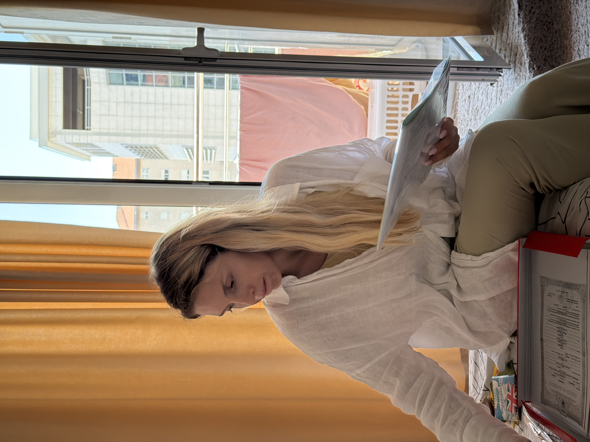 A woman with long blonde hair is sitting on a carpeted floor near a window. She is wearing a loose-fitting white shirt and light-colored pants. The woman is holding a document or book and appears to be reading it intently. Sunlight streams in through the window, illuminating her and the scene. Outside the window, a balcony with a laundry basket and some pink fabric is visible, as well as buildings in the background. The overall scene suggests a casual and relaxed moment, possibly during the daytime. On the floor next to the woman is a red folder containing what looks like official documents. A small item with the Union Jack flag is also visible on the floor.