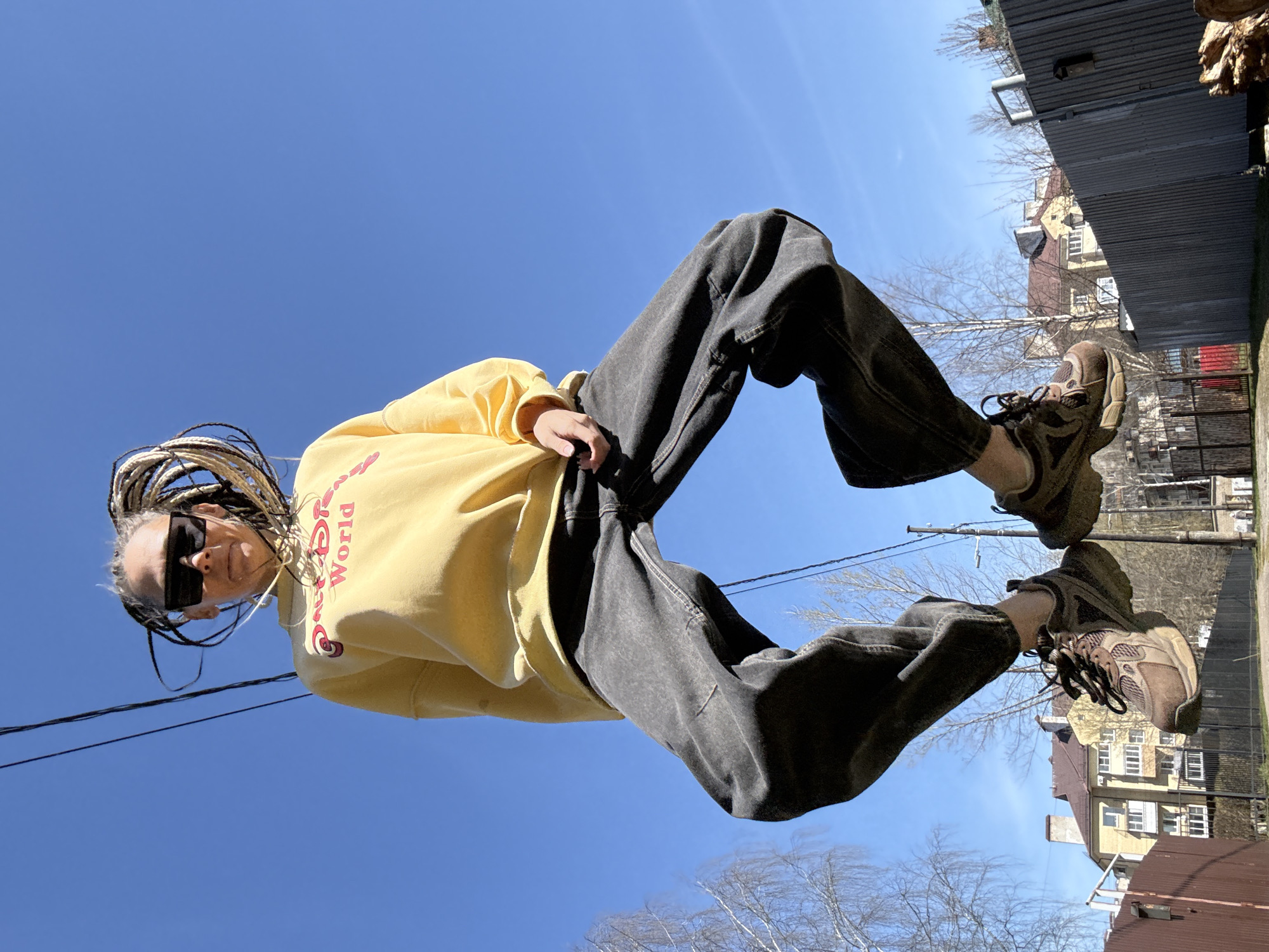 A person with dreadlocks wearing sunglasses, a yellow sweatshirt, and dark jeans is standing outside. The person is looking down at the camera. Behind the person is a clear blue sky. A building is visible in the background, as well as a fence. The photo appears to have been taken in the city of Bor, Russia.