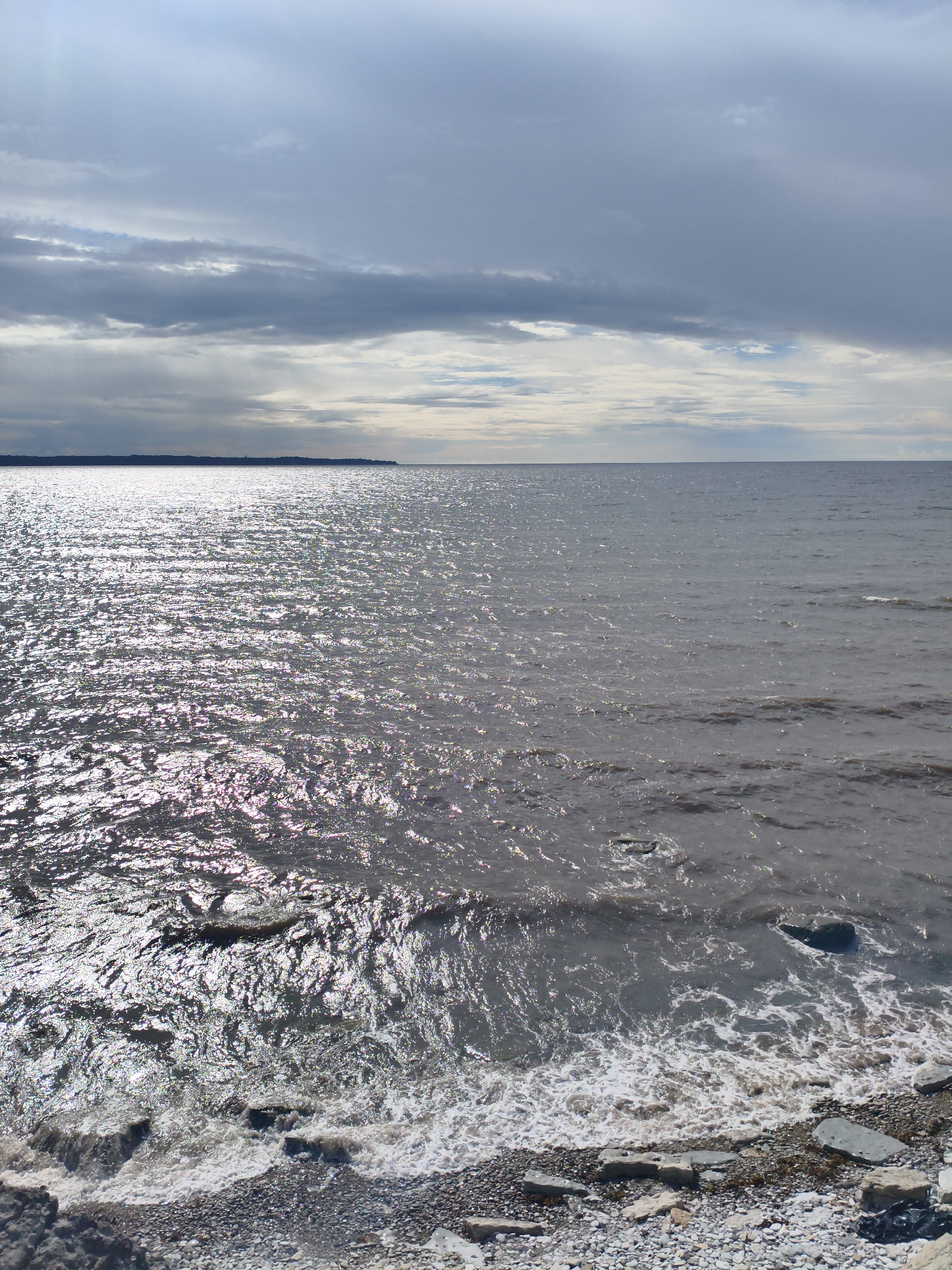 The image depicts a serene seascape on a cloudy day, likely near the coast of Paldiski, Estonia. The sky is filled with a mix of dark, heavy clouds and brighter, lighter patches, suggesting an overcast atmosphere with some sunlight breaking through. The sea is calm, with gentle waves lapping against a pebbled shore dotted with flat, grey stones. The water reflects the sunlight, creating a shimmering effect that stretches across the surface. In the distance, a faint landmass is visible on the horizon, with a subtle outline of trees. The overall impression is one of a quiet, natural landscape. There are no people or animals present in the image. The scene is static and peaceful, emphasizing the vastness of the sea and sky.