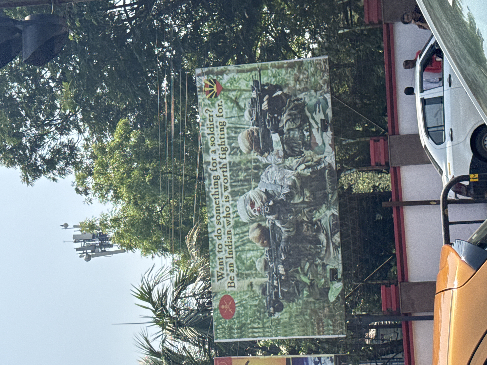 The image captures a street scene in Kolkata, India, featuring a large advertisement for Indian Army recruitment. The billboard displays a group of soldiers in camouflage gear and reads, "Want to do something for a soldier? Be an Indian who is worth fighting for." The backdrop of the billboard suggests a jungle or forested environment. A white van and an orange car are partially visible near the billboard, indicating road traffic. The sky is visible in the upper portions, partly obscured by trees and communication infrastructure.