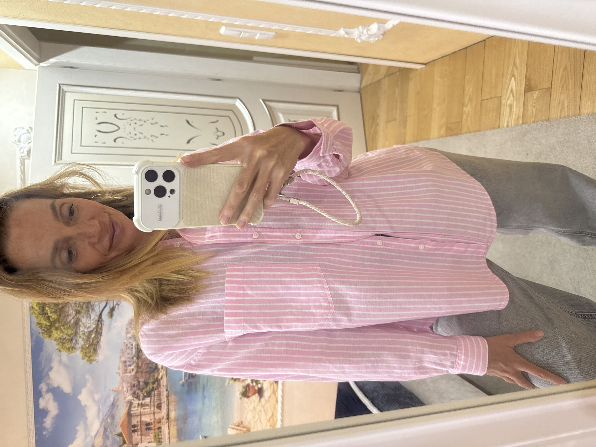 A woman is taking a mirror selfie in what appears to be her home. She is wearing a pink and white striped button-down shirt and gray pants. She is holding an iPhone in her right hand. The background shows a door with a decorative design, wooden flooring, and a seascape print on the mirror, portraying buildings along the coast and water, resembling a Mediterranean or European coastal city. There is no evidence to suggest the photo was taken in Saint Petersburg, Russia.