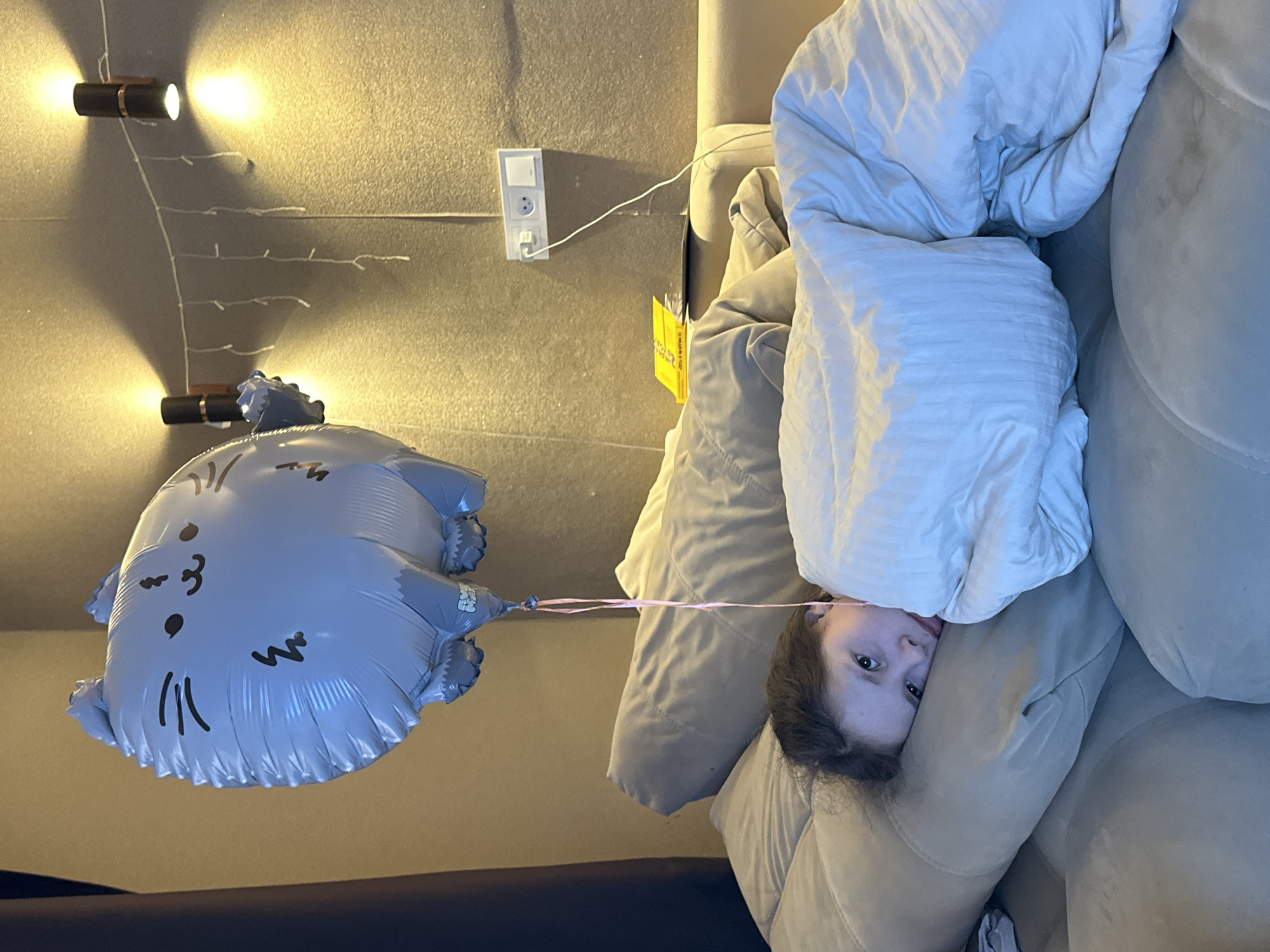 A child is partially visible, lying face-up on a light brown couch with a white comforter partially covering their face. A gray, cat-shaped balloon is held by a pink string connected to the child. The background includes a light brown wall with two mounted lights and a white electrical outlet. A yellow paper or card is visible near the couch. The location appears to be indoors, in an unknown city and state.