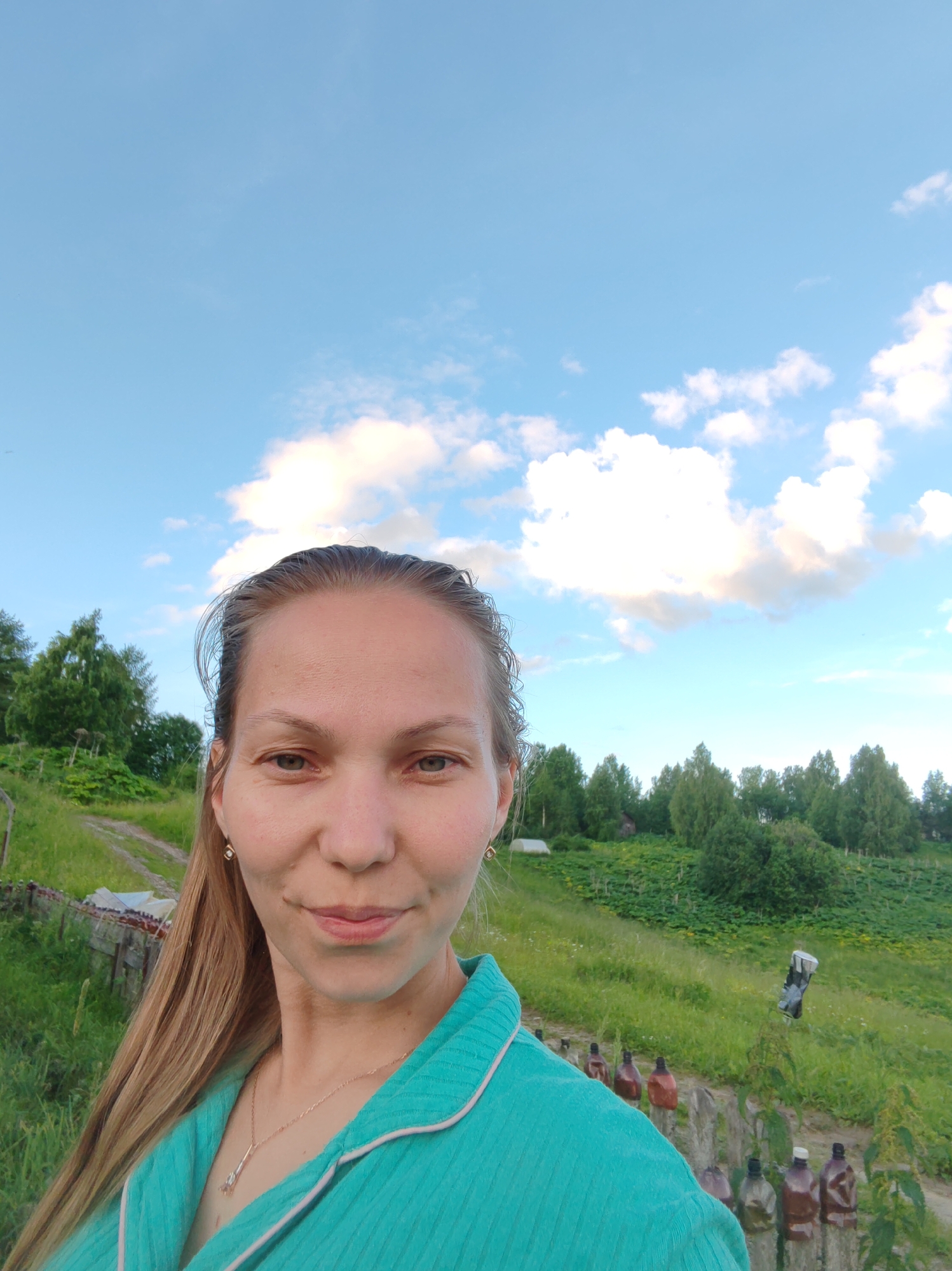 A woman is taking a selfie outdoors with a bright blue sky and fluffy white clouds overhead. She has long blonde hair pulled back and is wearing a teal robe with white trim. A delicate gold necklace with a crystal pendant is visible. The background depicts a lush green landscape with rolling hills, trees, and a dirt path. There are several wooden posts with various colored bottles on them, resembling a rustic fence or decoration. In the distance, a figure that appears to be a scarecrow stands on a hillside. The lighting suggests it is daytime, possibly late afternoon or early evening, with soft light illuminating the scene. The overall mood is relaxed and natural, hinting at a peaceful rural setting. There is no visible text in the image.