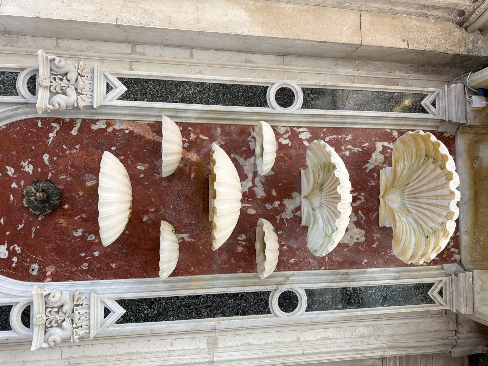 This is a medium shot, high-angle view of a decorative marble wall in Saint Petersburg, Russia. The wall features a red marble panel inset with a black granite band, bordered by white marble. The red marble panel is adorned with several carved white marble shell-like objects, varying in size and depth, arranged horizontally across the panel. To the far left of the red marble is a dark, ornate metal rosette.
The white marble framing the red panel includes decorative carved elements. On the left and right edges of the black granite band are triangular white marble accents. Above and below the red marble panel are white marble sections with carved decorative elements that include what appear to be two coiled serpents intertwined with floral motifs and a crown-like structure. Circular decorative elements are also present within the white marble framework. The overall impression is one of opulent architectural decoration. There is no discernible text in the image. The lighting suggests an indoor setting, possibly a church or a grand building, with no indication of time of day or weather. The scene depicts static decorative elements rather than an active event.