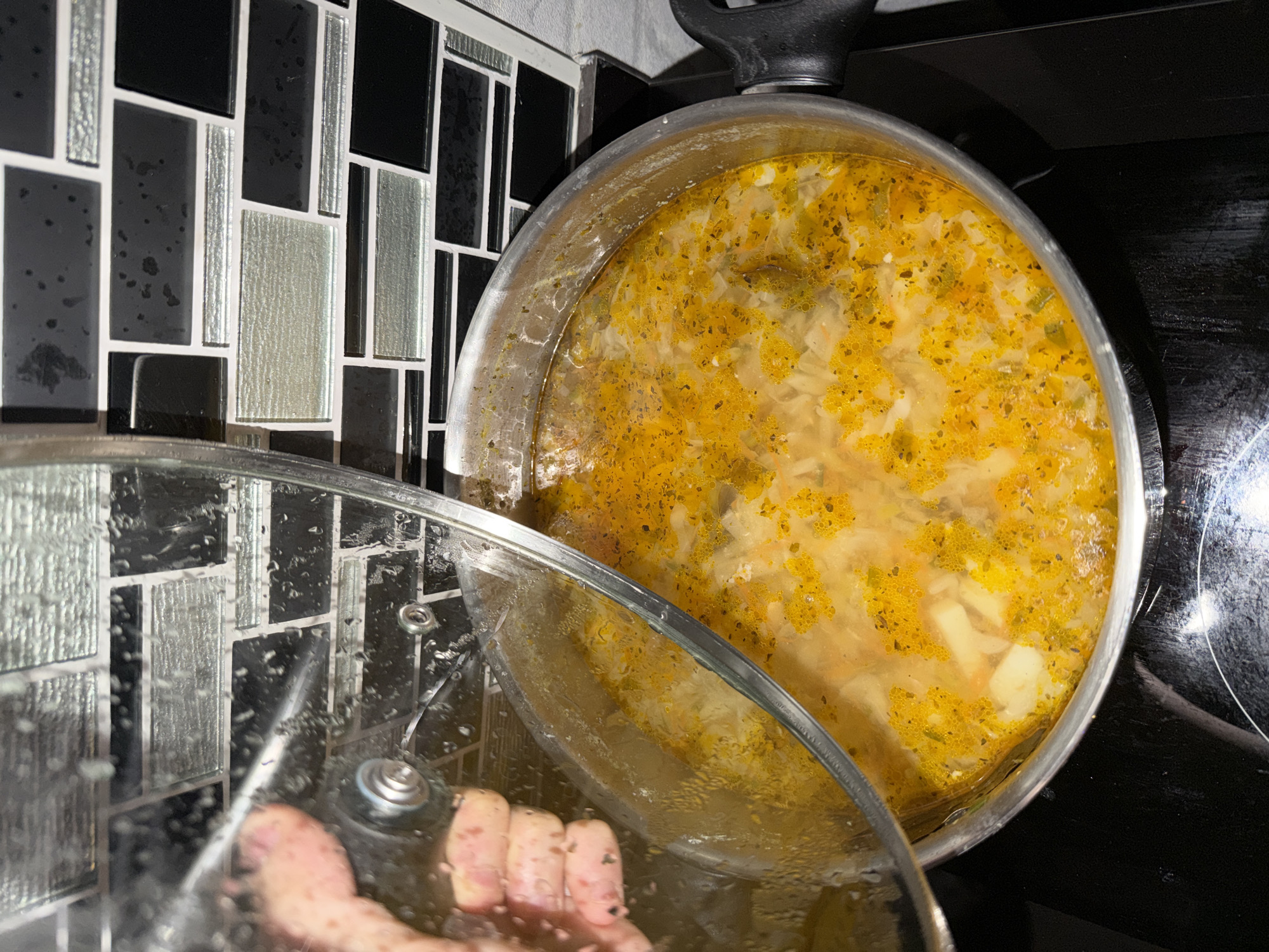 A hand lifts a glass lid off a pot of simmering soup. The soup is a vibrant yellow-orange and appears to contain chunks of vegetables and possibly noodles. The pot is on a black stovetop, and the background features a tiled backsplash with black and grey rectangular tiles. The scene is well-lit, suggesting an indoor setting. There is no indication of the specific location within Bangkok, Thailand, or any other city.